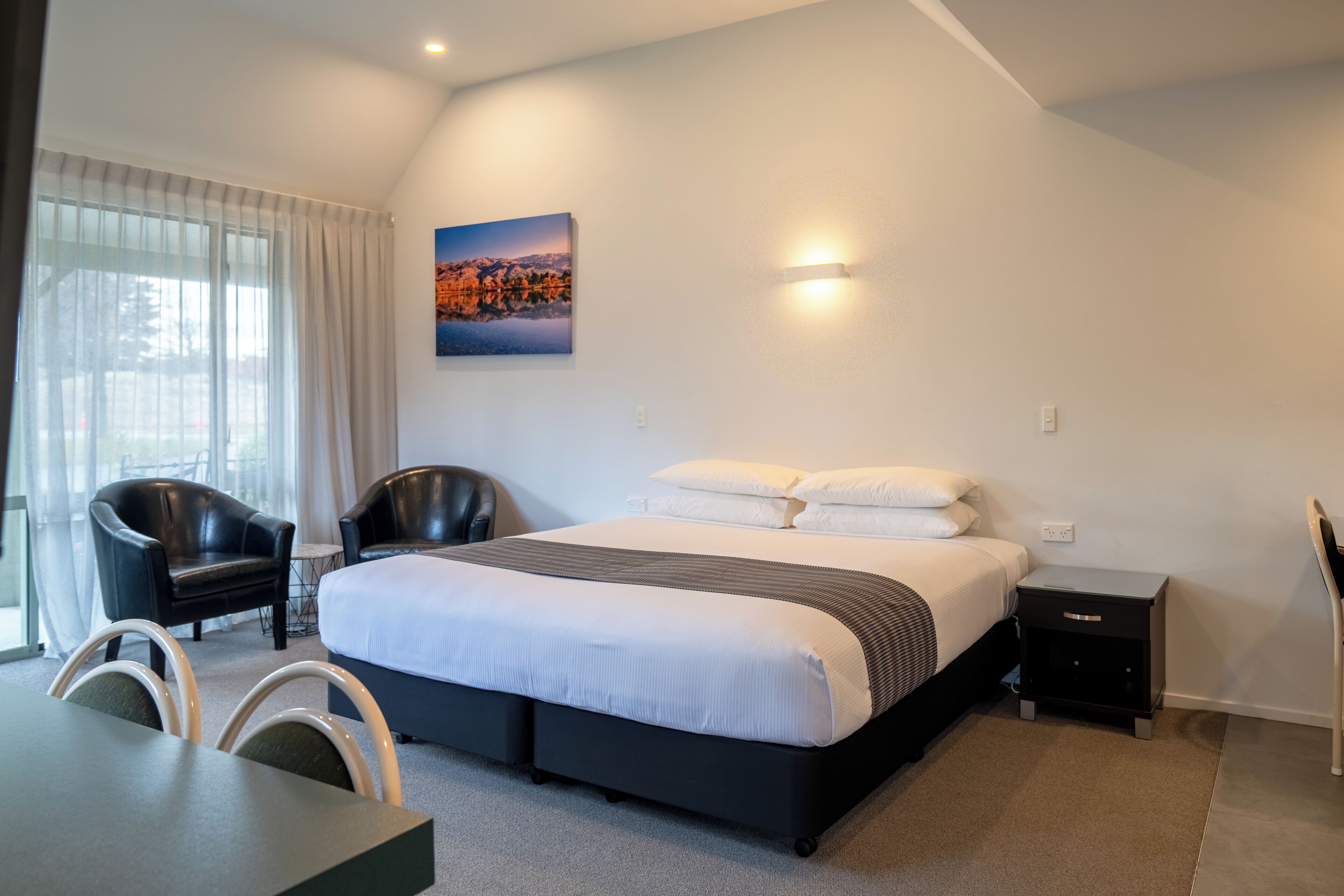 A modern hotel room with a large bed, two black chairs, a small table, and a painting on the wall.