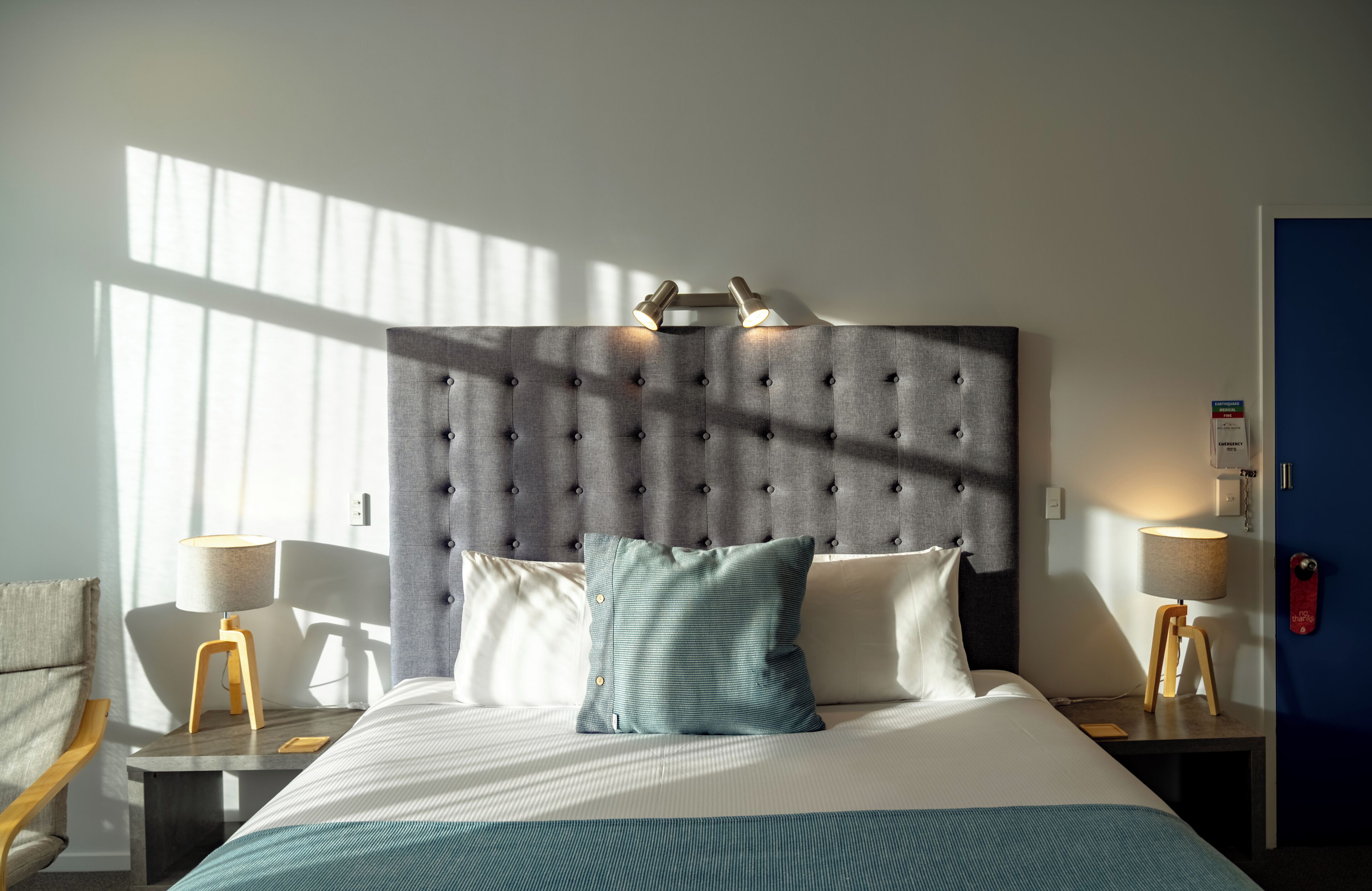 Een hotelkamer met een gestoffeerd hoofdeinde, een bed met wit en teal beddengoed, twee nachtkastjes met lampen, en een uitzicht buiten door een raam.