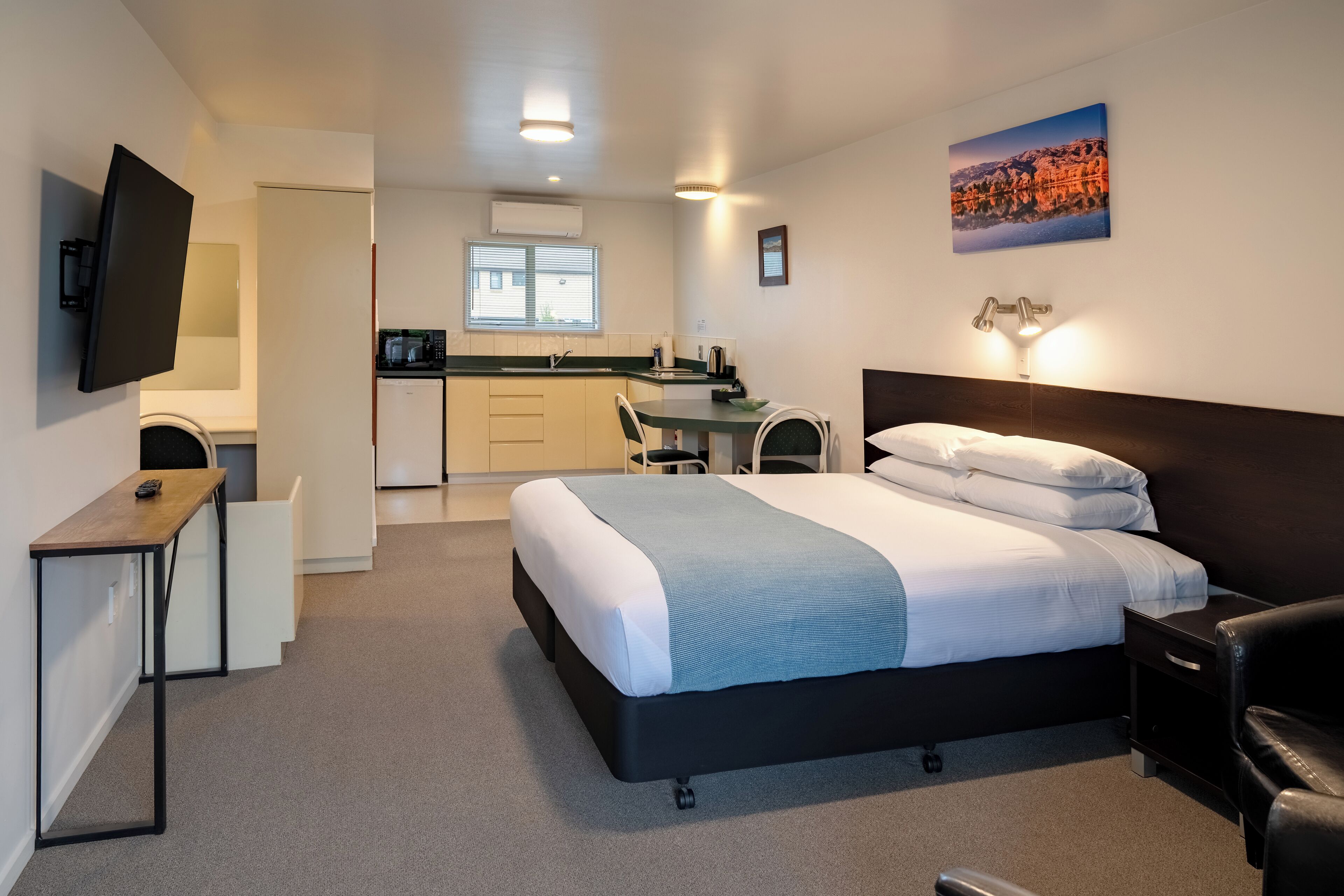 A hotel room with a bed, a desk, a chair, a TV, and a kitchenette in the background.