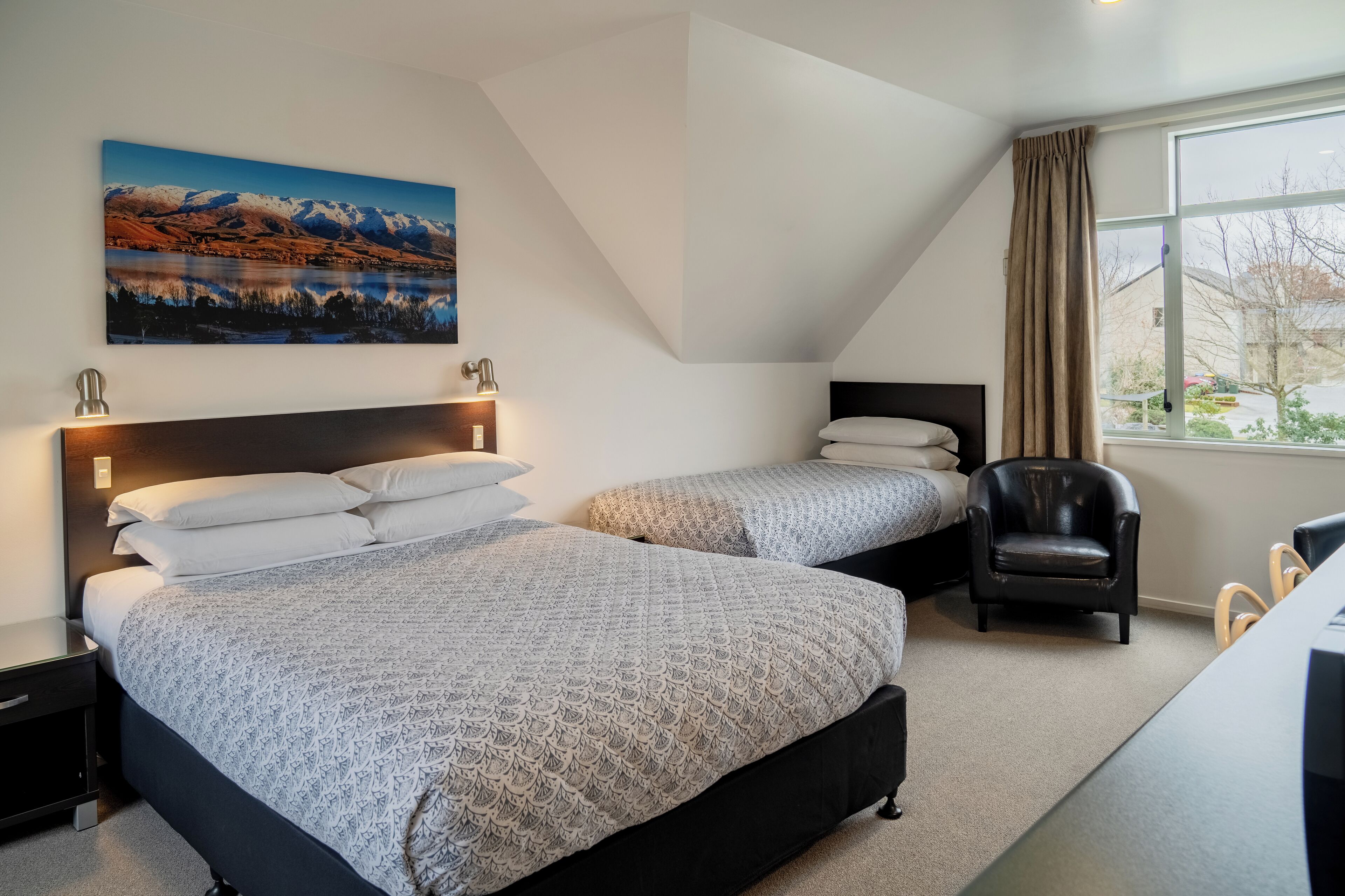 Une chambre d’hôtel avec deux lits, un bureau, une chaise et une grande fenêtre donnant sur les montagnes et un lac.