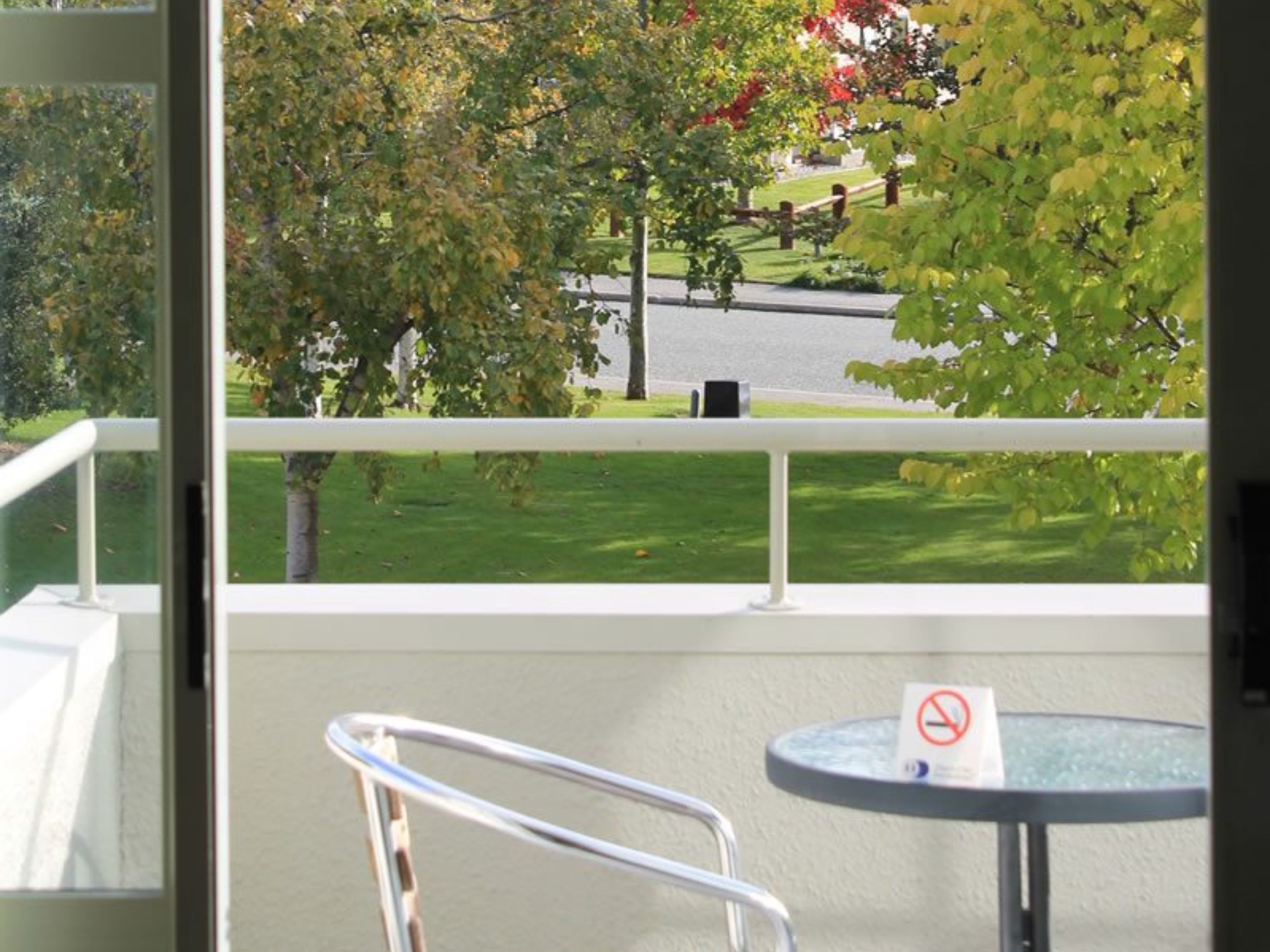 Un balcone con vista su un prato verde e alberi, una ringhiera bianca e un tavolo rotondo con un cartello che indica il divieto di fumare.