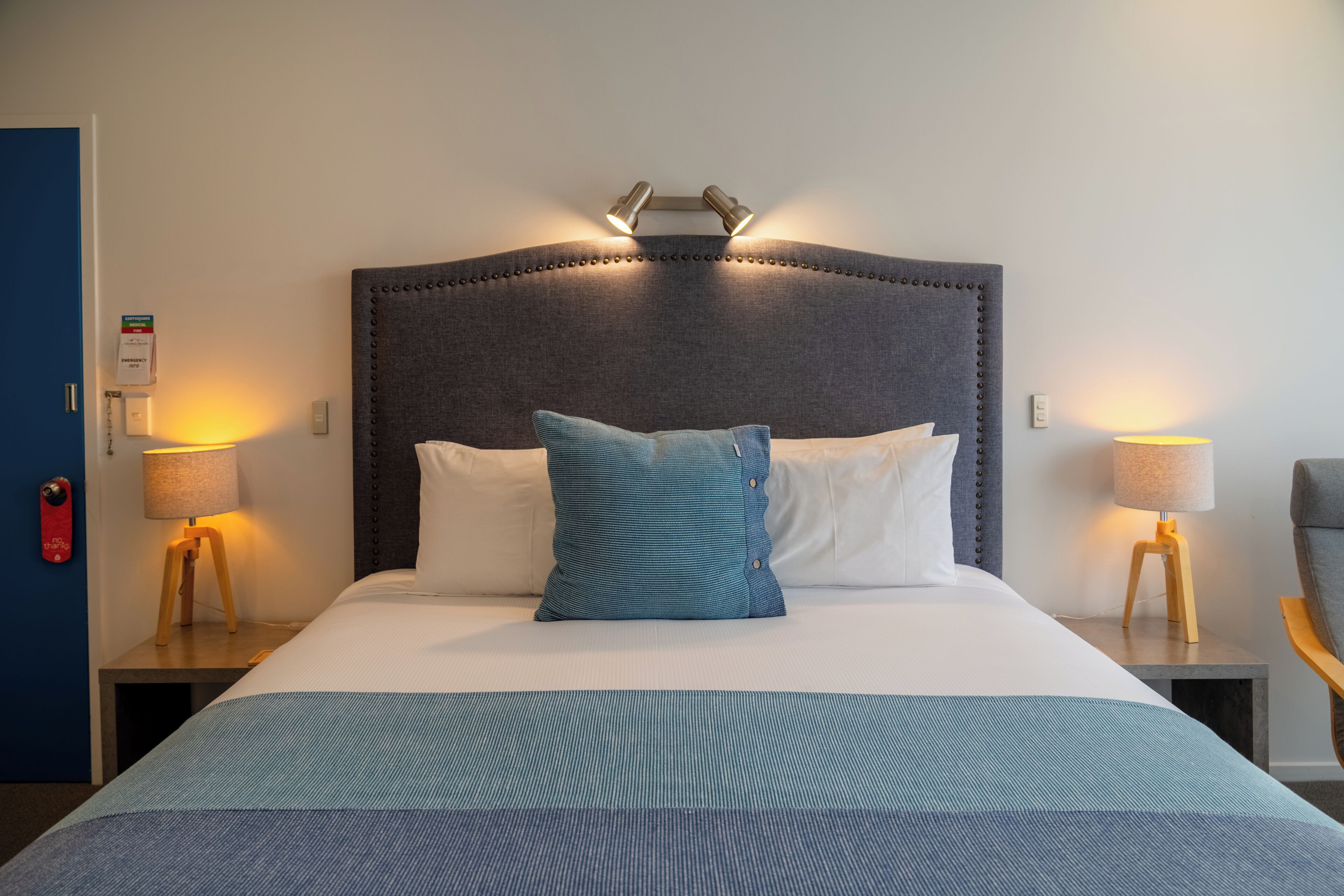 A hotel room with a large bed, two bedside tables with lamps, a blue door, and a wall-mounted light fixture above the bed.