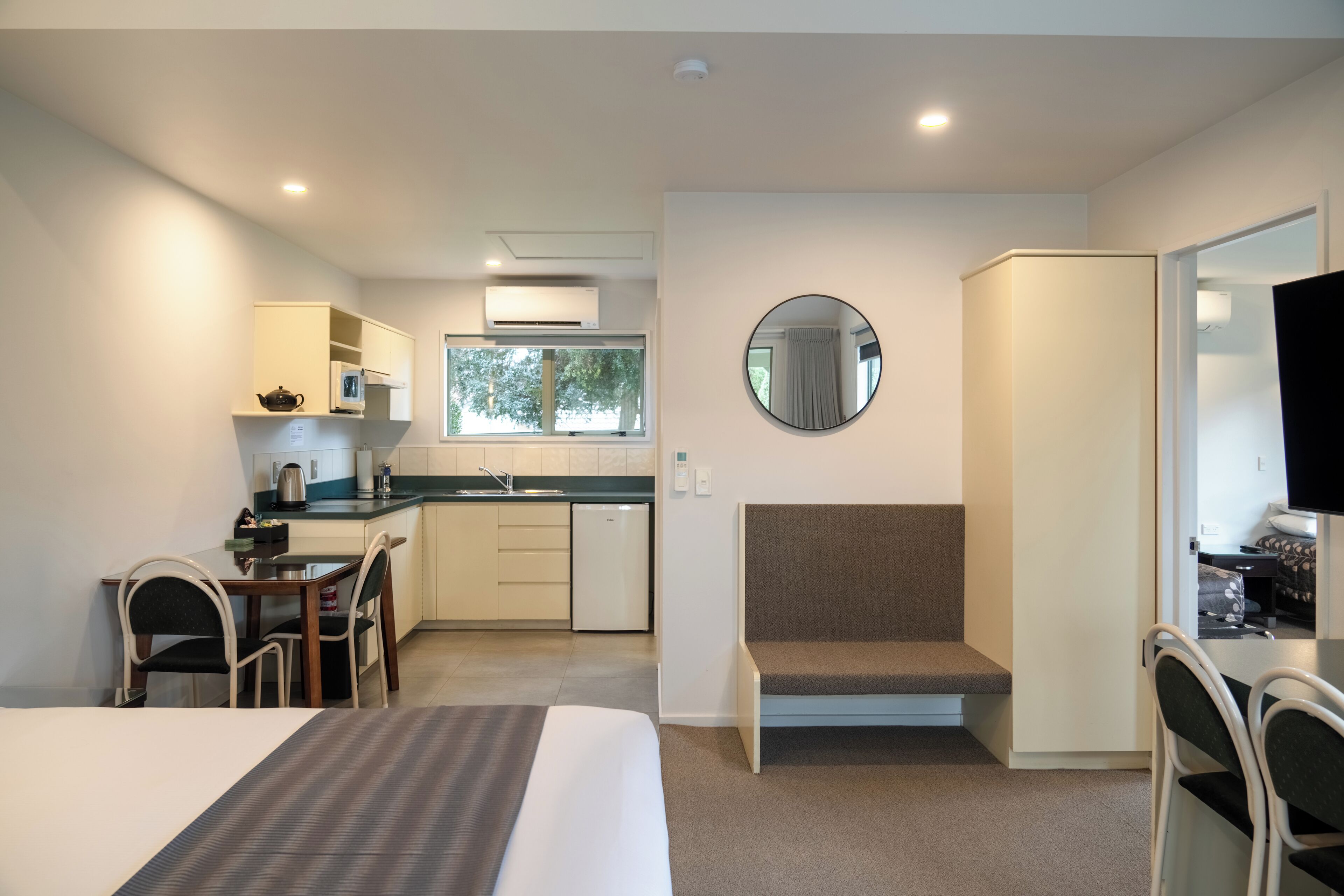 A compact hotel room with a bed, a small kitchenette, a dining area, and a seating area with a mirror.