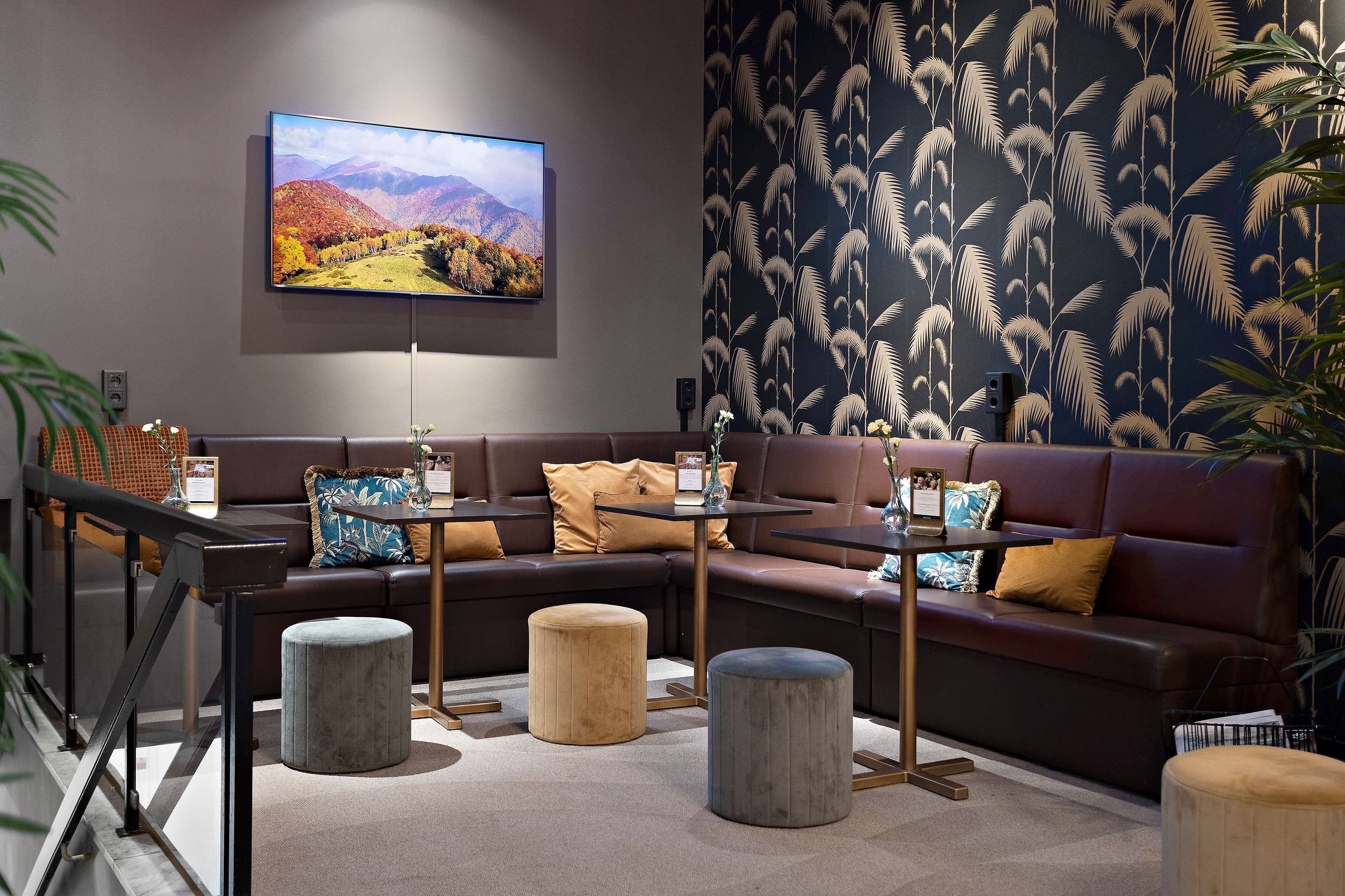 Ein moderner Lounge-Bereich mit einem großen Flachbildfernseher an der Wand, einem Wandbild mit Muster, einem braunen Ledersofa mit Kissen und einem kleinen runden Tisch mit einer Vase.