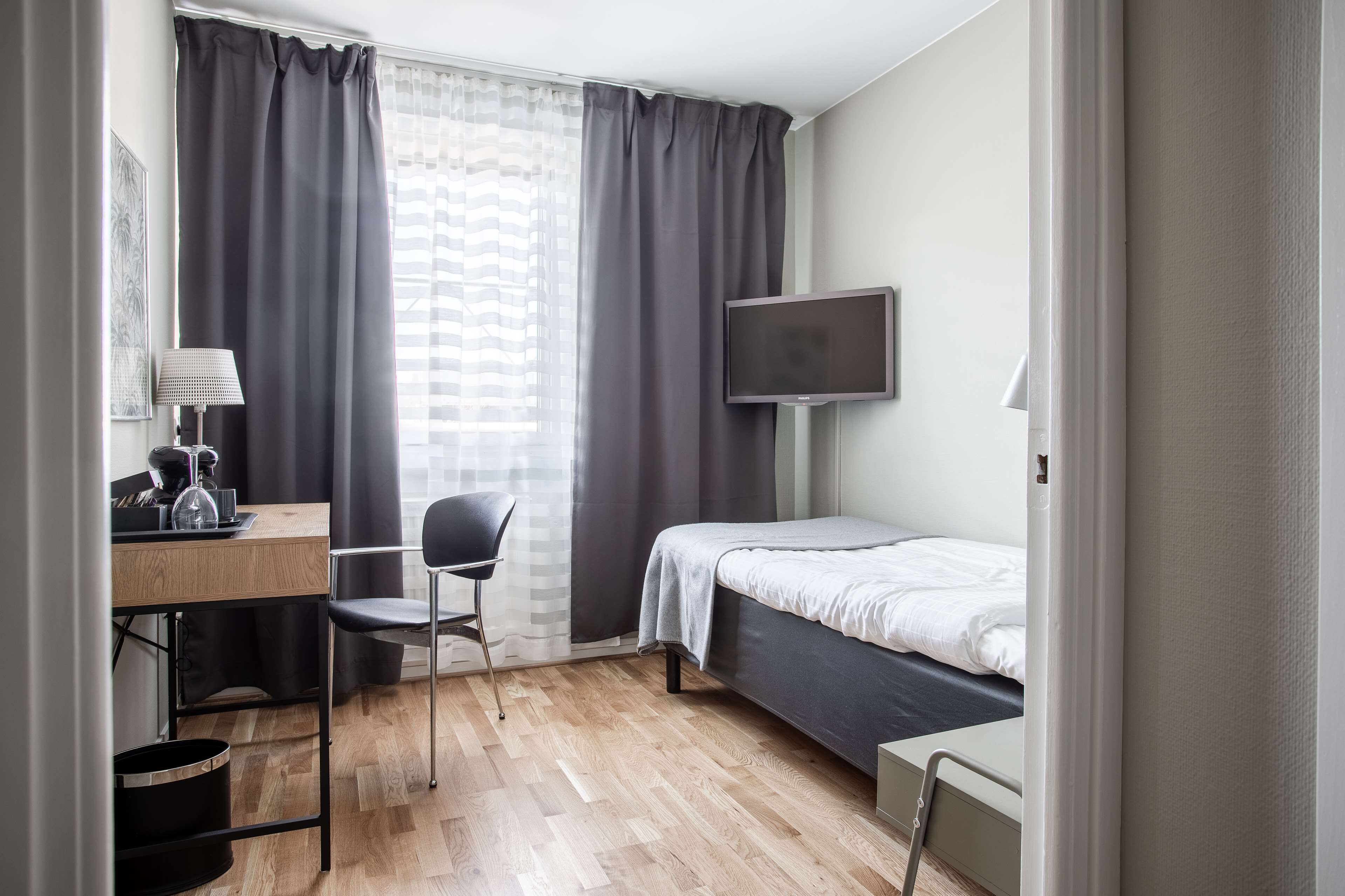A compact hotel room with a bed, a desk with a chair, a TV, and a window with curtains.