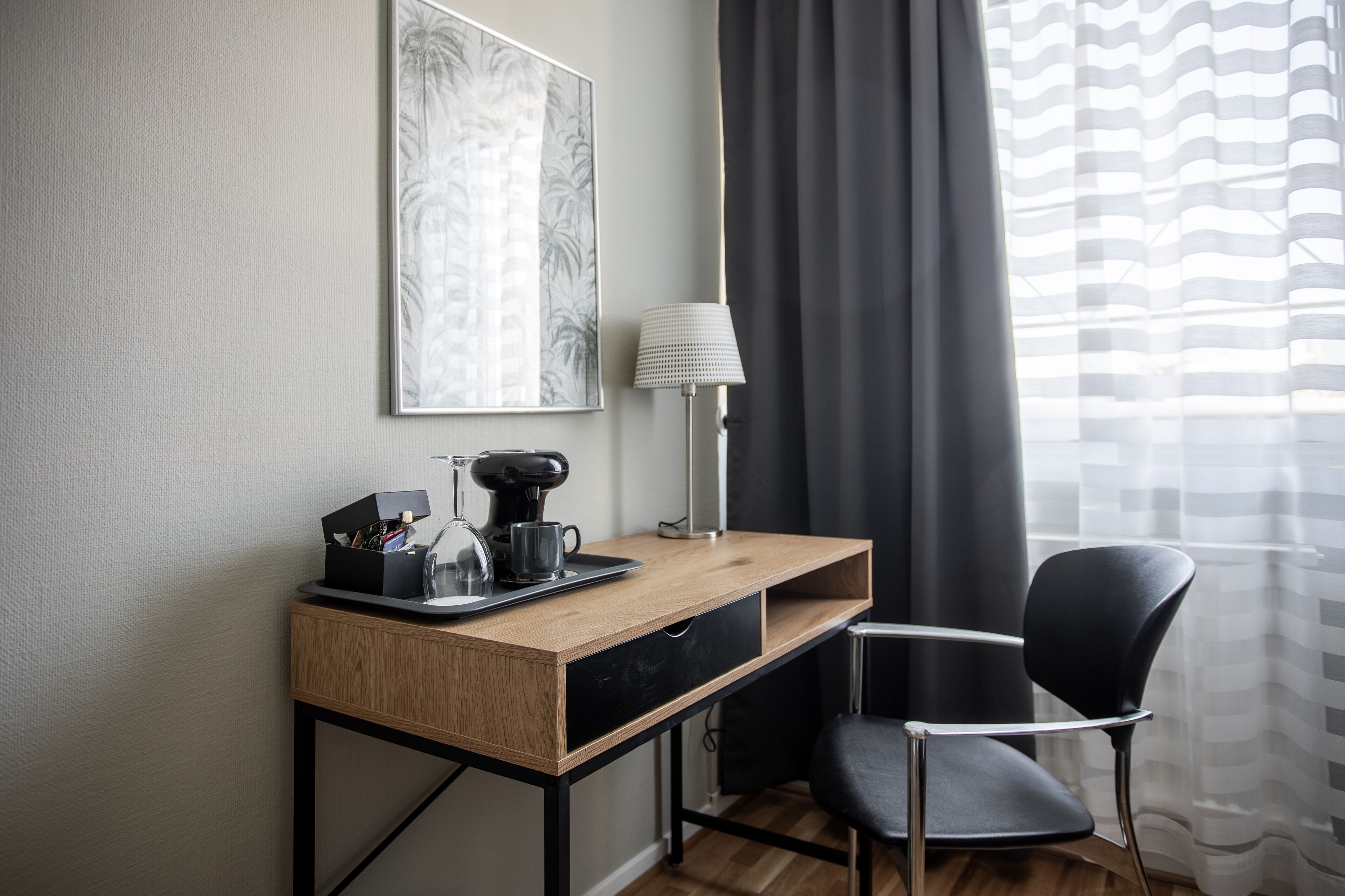 Una habitació d'hotel amb escriptori de fusta, cadira negra, cafetera, làmpada i un quadre amb marc a la paret.