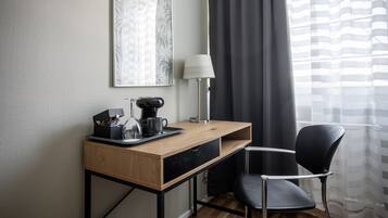 Una habitació d'hotel amb escriptori de fusta, cadira negra, cafetera, làmpada i un quadre amb marc a la paret.
