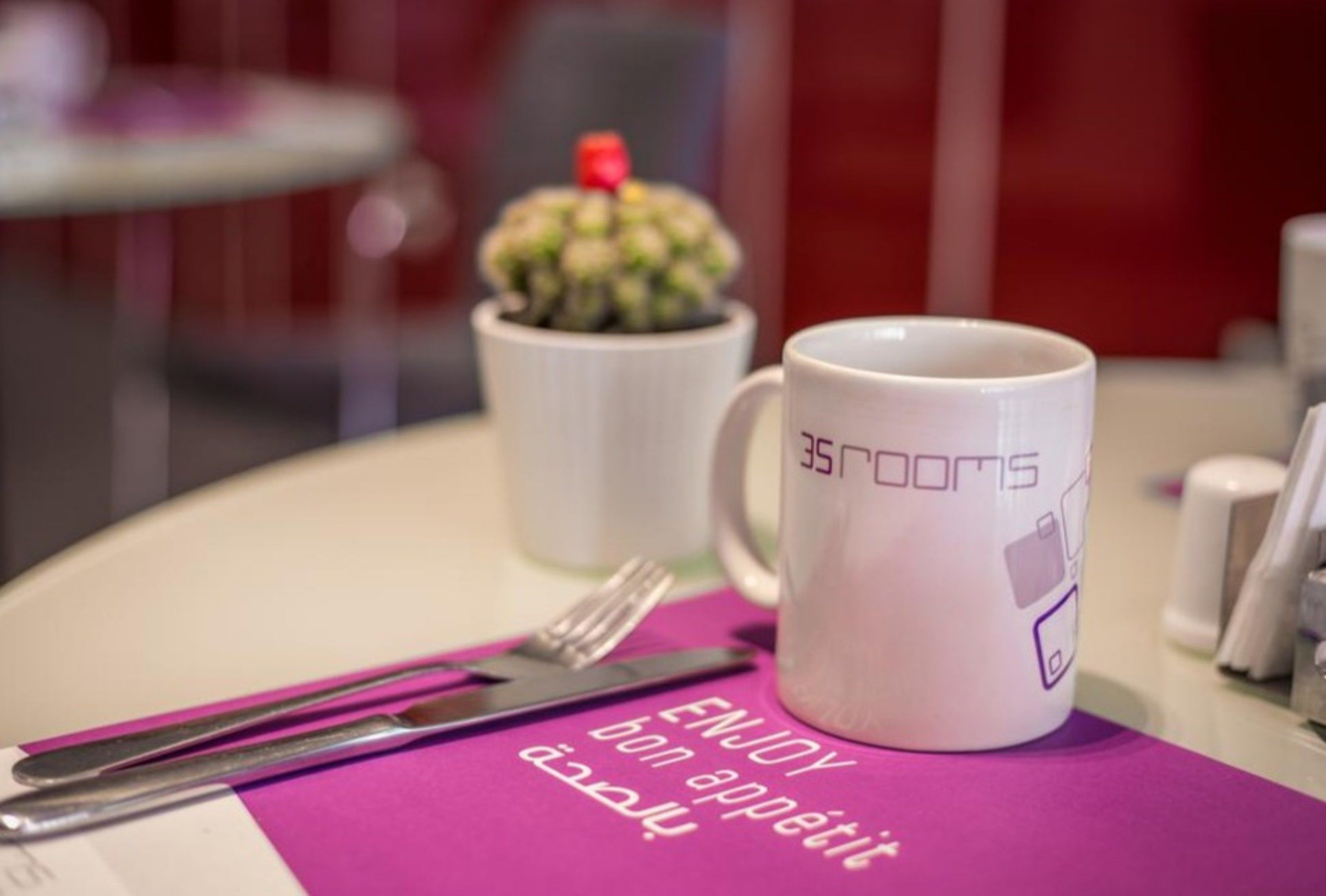 A table with a white mug labeled "35ROOMS" and a fork on a purple placemat with text.
