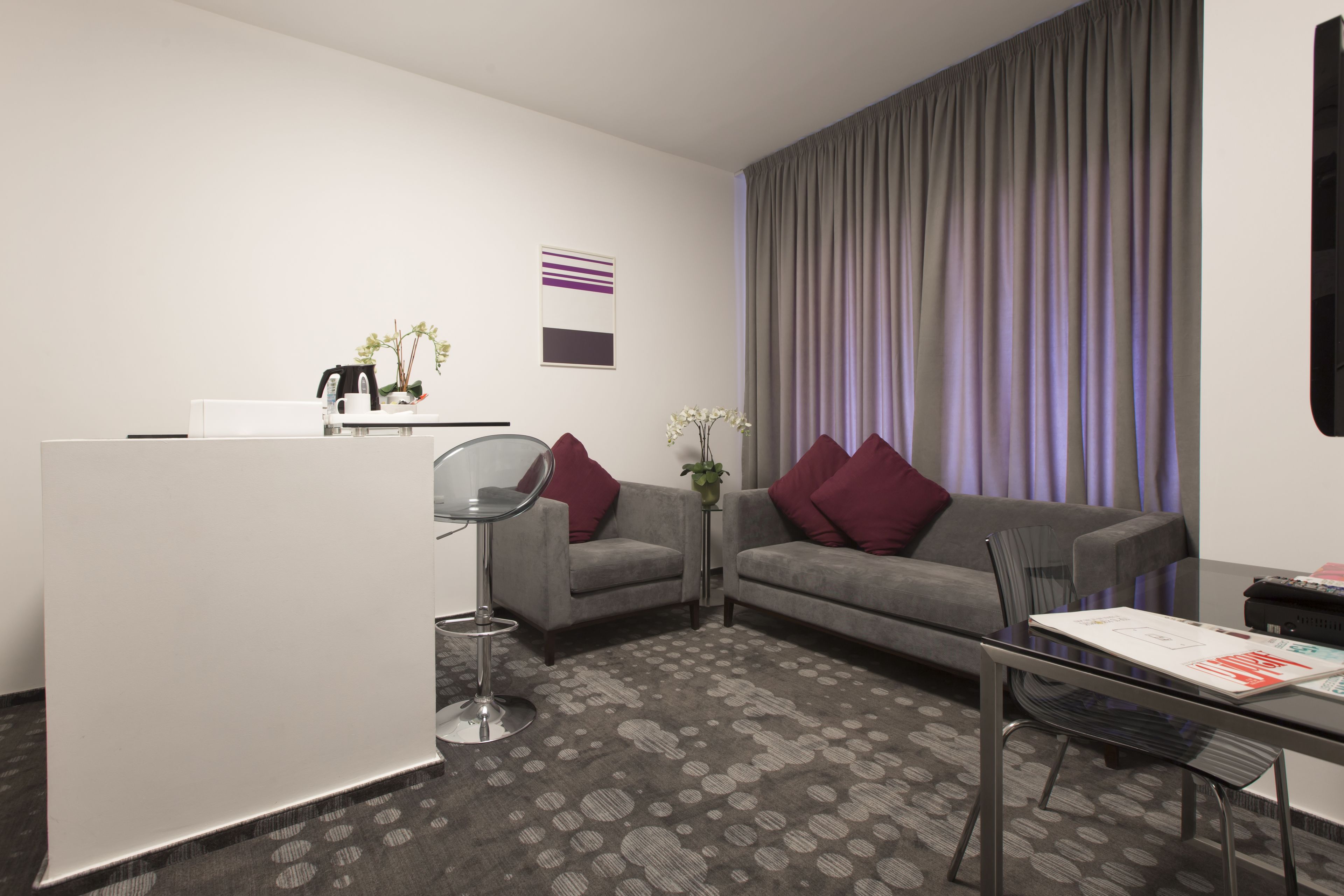 Ett modernt hotellrum med en grå soffa, ett litet sidobord med en vas med blommor, ett glasbord och ett stort fönster med gardiner.