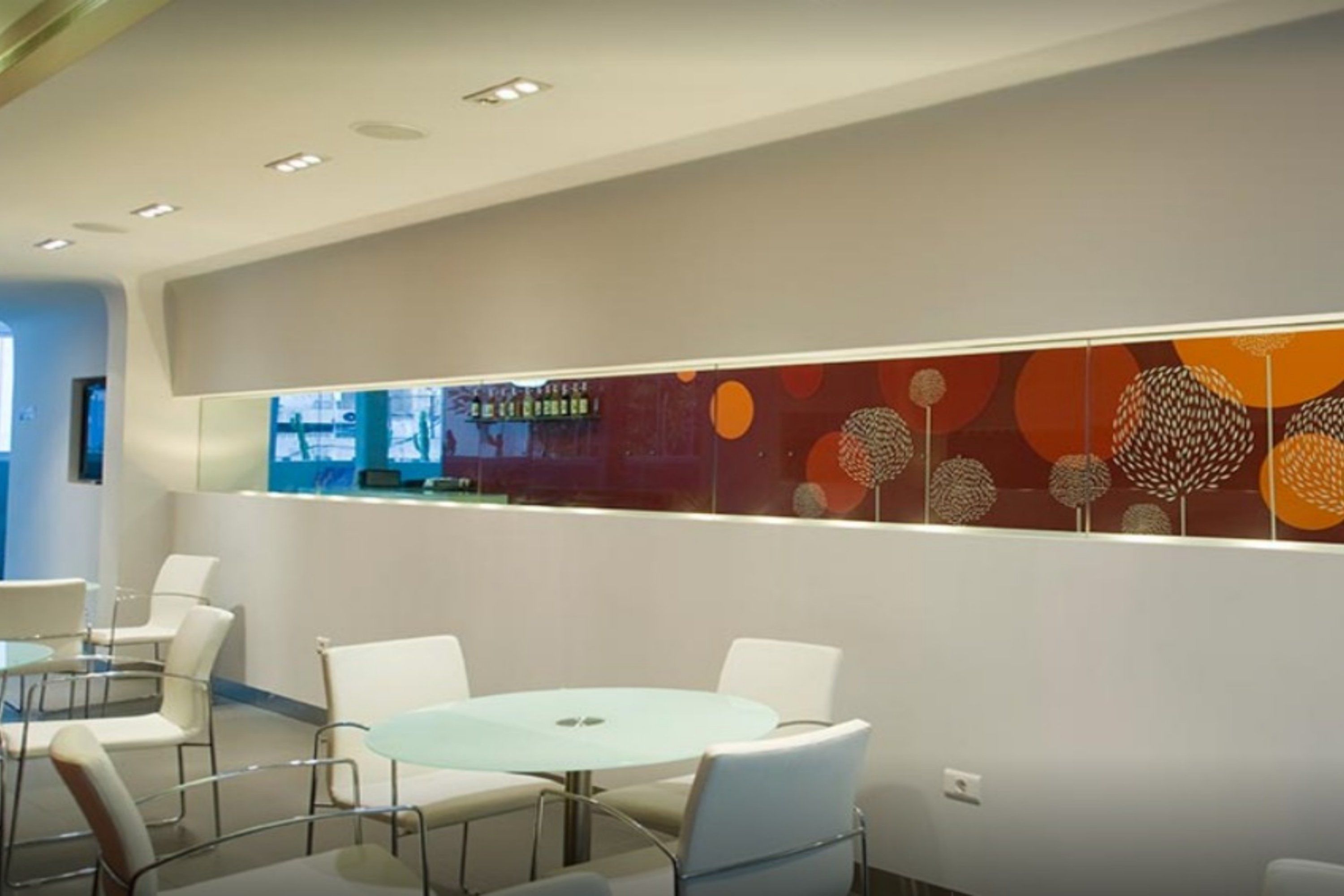 A modern interior with a round glass table and white chairs, a decorative wall with circular patterns, and recessed ceiling lights.