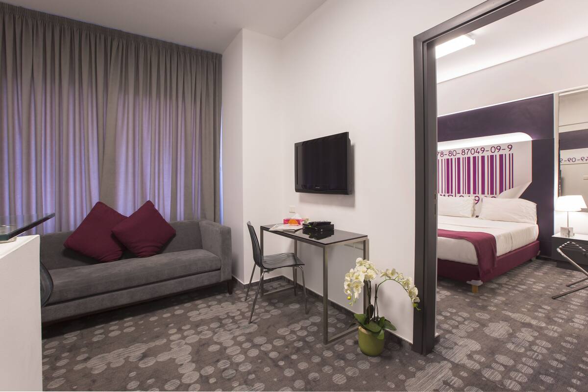 Ein Hotelzimmer mit grauem Sofa, einem kleinen Tisch, einem Fernseher, einem Bett mit weißer und violetter Bettdecke sowie einer Zimmerpflanze mit Orchideen.