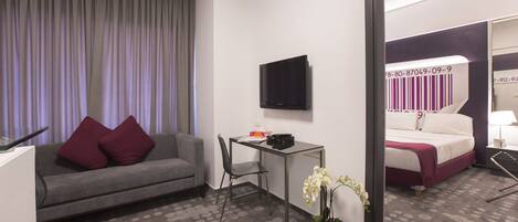 A hotel room with a grey sofa, a small table, a TV, a bed with a white and purple bedspread, and a potted orchid.