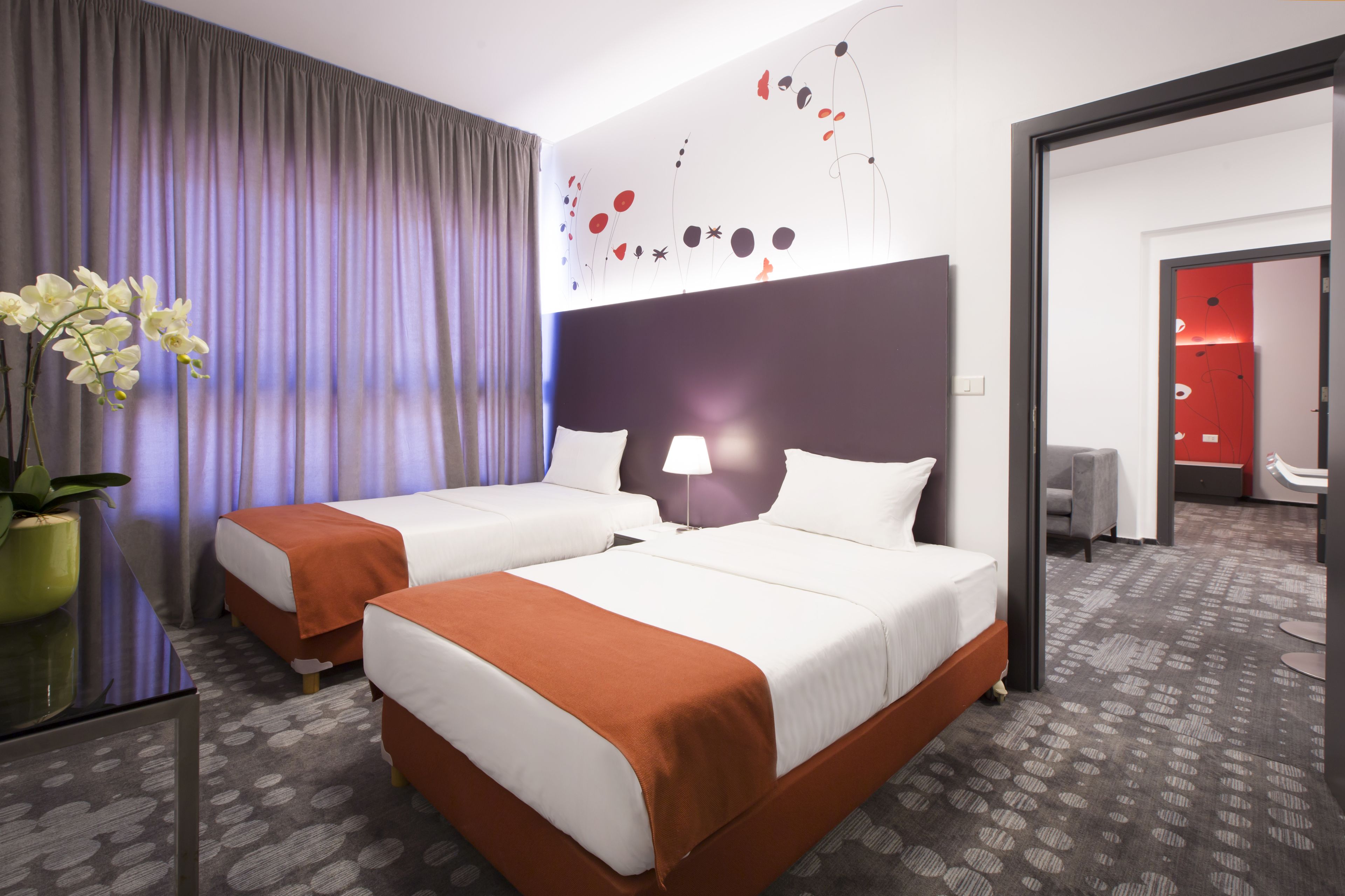 A hotel room with two beds, a large window with curtains, a wall art piece, and a potted plant.