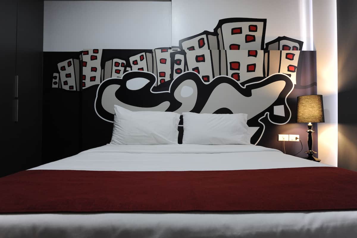 Ein Bett mit weißer und roter Bettdecke, einem schwarz-weißen Kopfteil und einer Wand mit Stadtansicht-Mural.