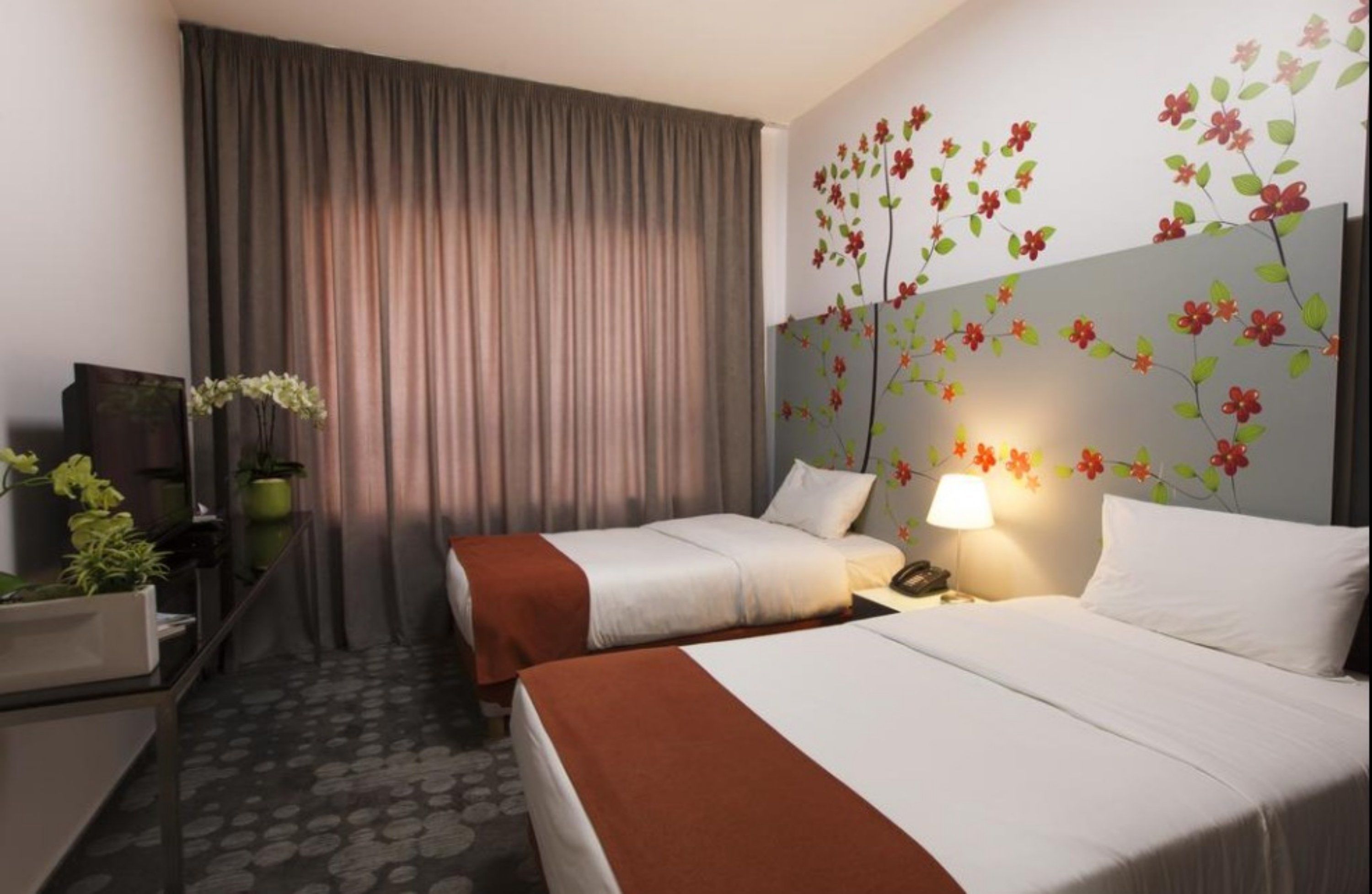 Habitación de hotel con dos camas, un ventanal con cortinas, una pared con una obra de arte floral y un escritorio con lámpara y teléfono.