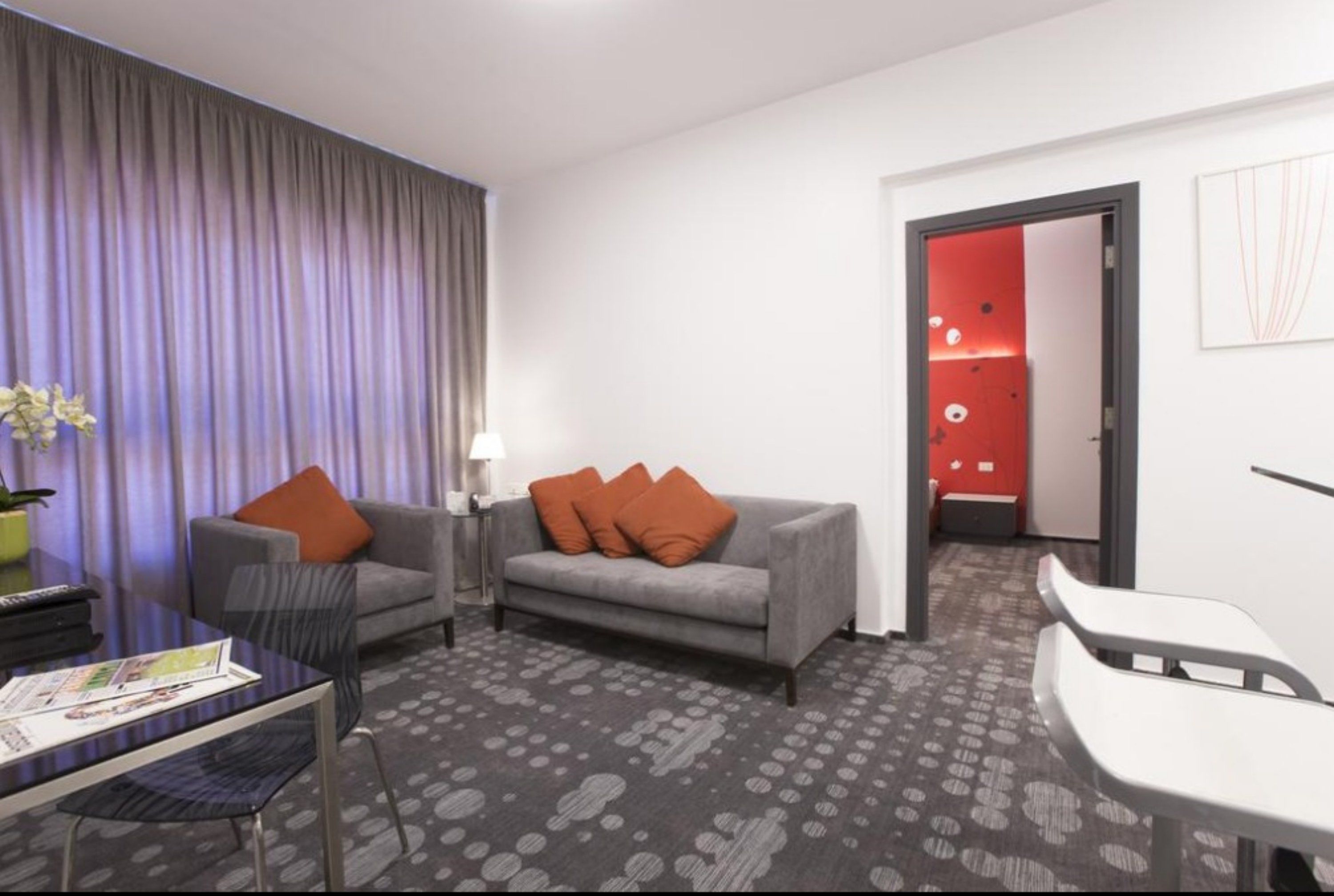 Une chambre d’hôtel moderne avec un canapé gris, des coussins orange, une table basse en verre, une lampe et un tapis à motifs.