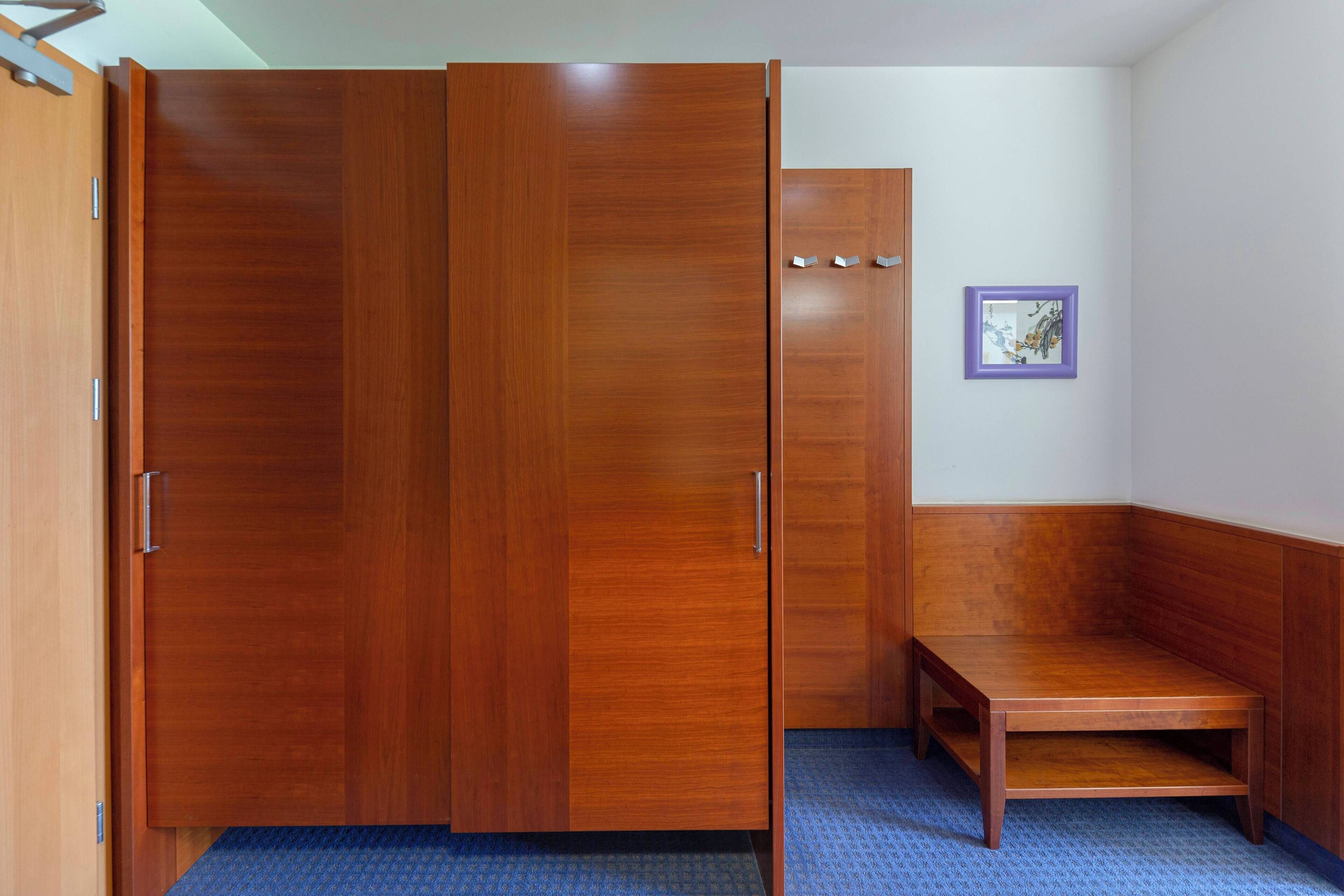 A room with a wooden wardrobe, a small bench, and a framed picture on the wall.