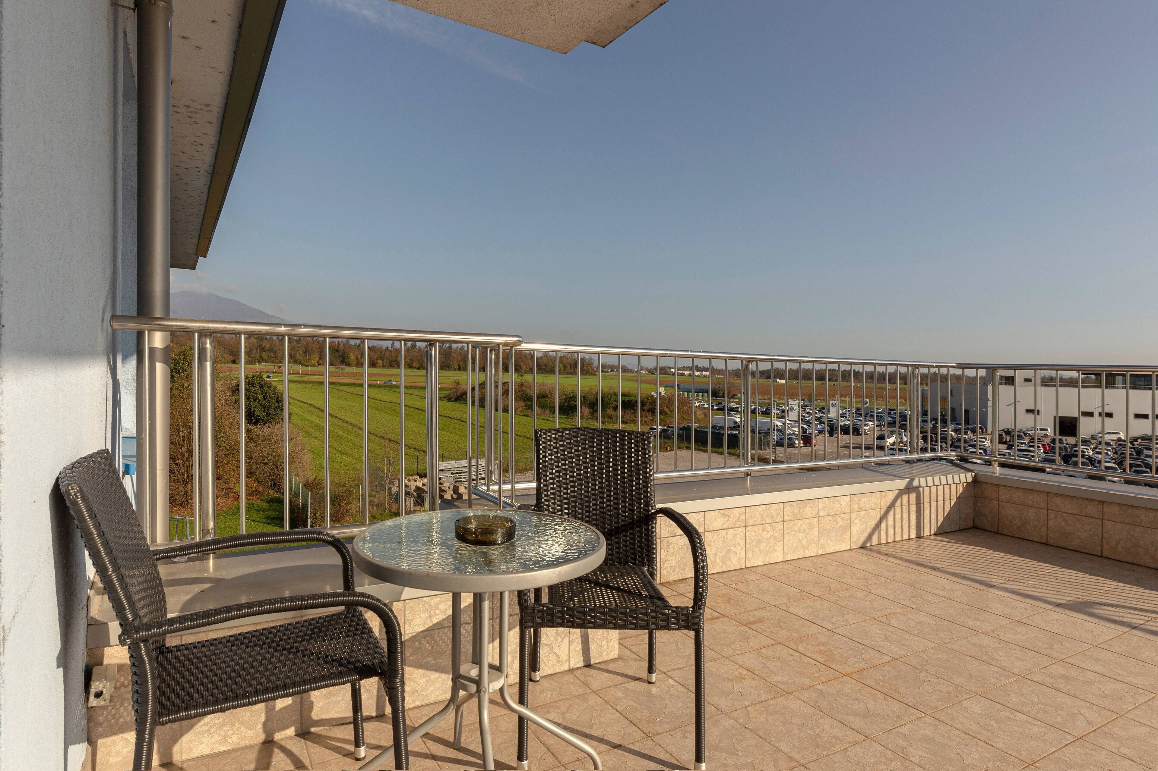 A balcony with two wicker chairs and a small round table, overlooking a parking area and a green field.