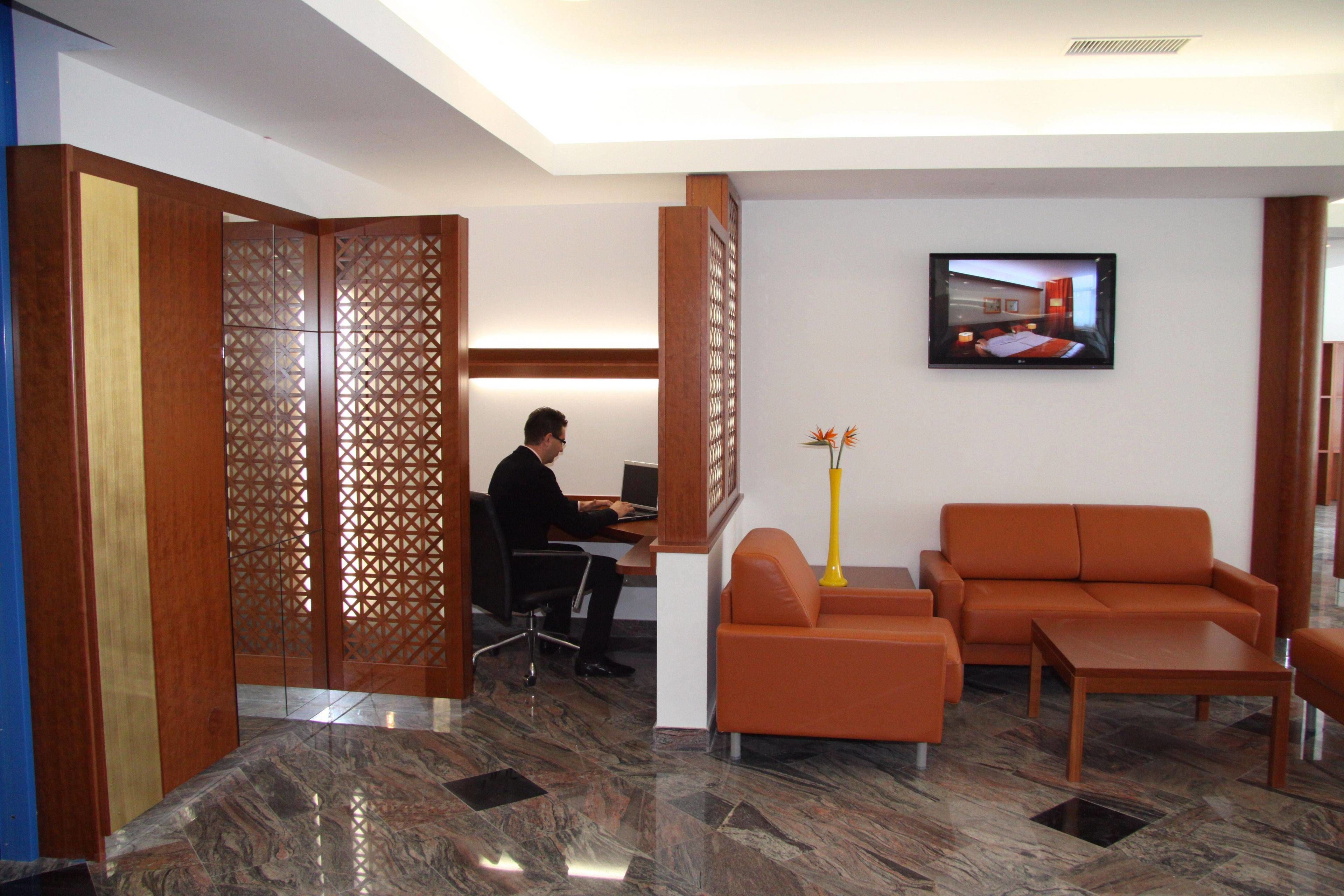 A modern hotel lobby with a reception desk, a seating area with orange sofas, and a TV mounted on the wall.