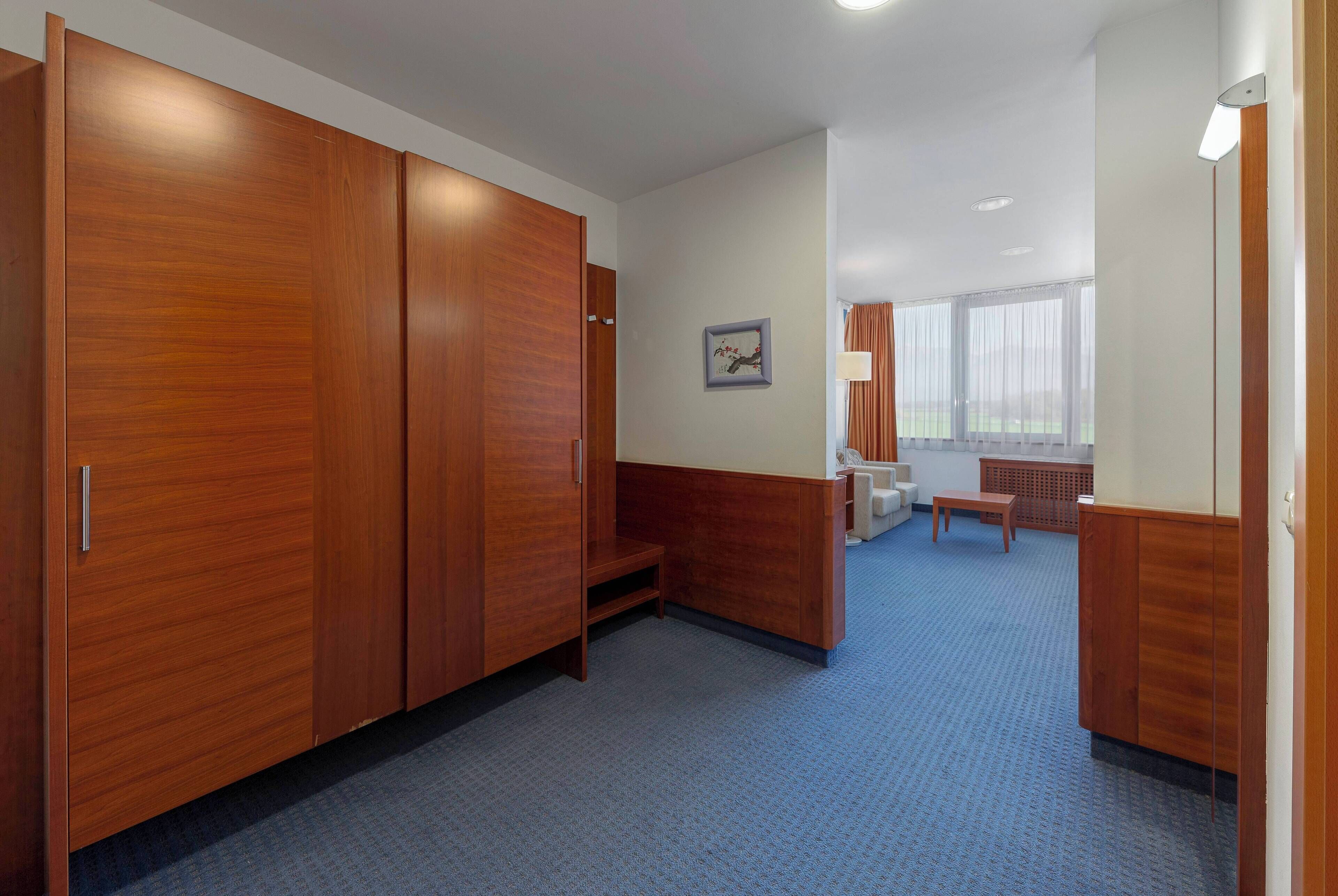 A hotel room with a large wooden wardrobe, a small seating area, and a window with curtains.