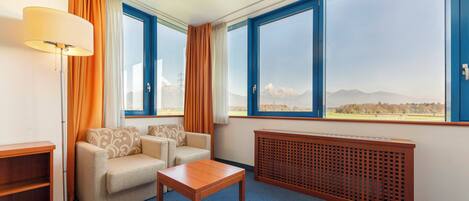 A hotel room with a sofa, a coffee table, a floor lamp, a radiator, and large windows with a mountain view.