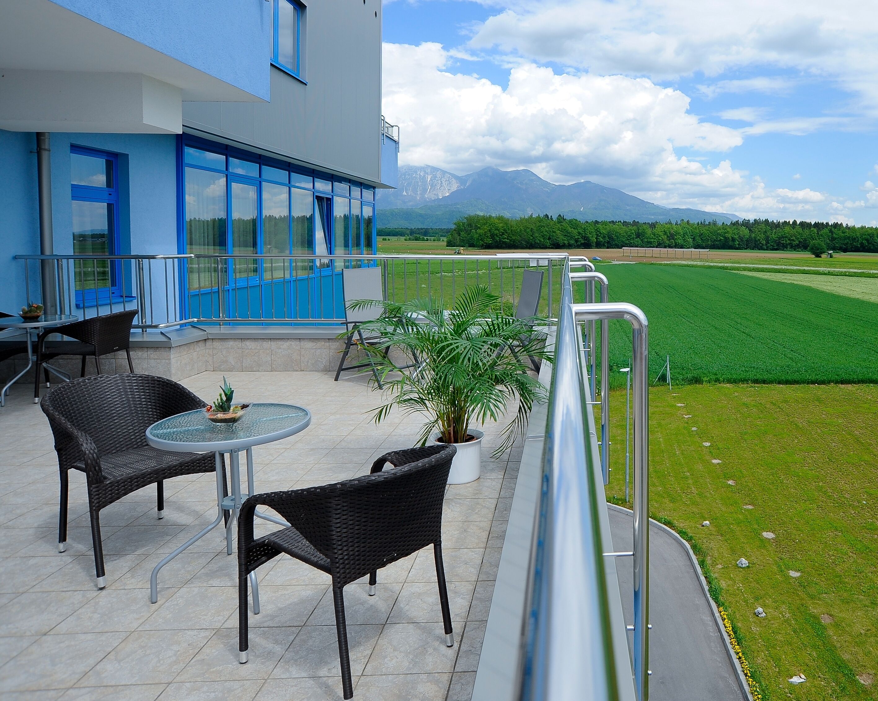 A balcony with wicker chairs and a table, overlooking a green field and mountains.