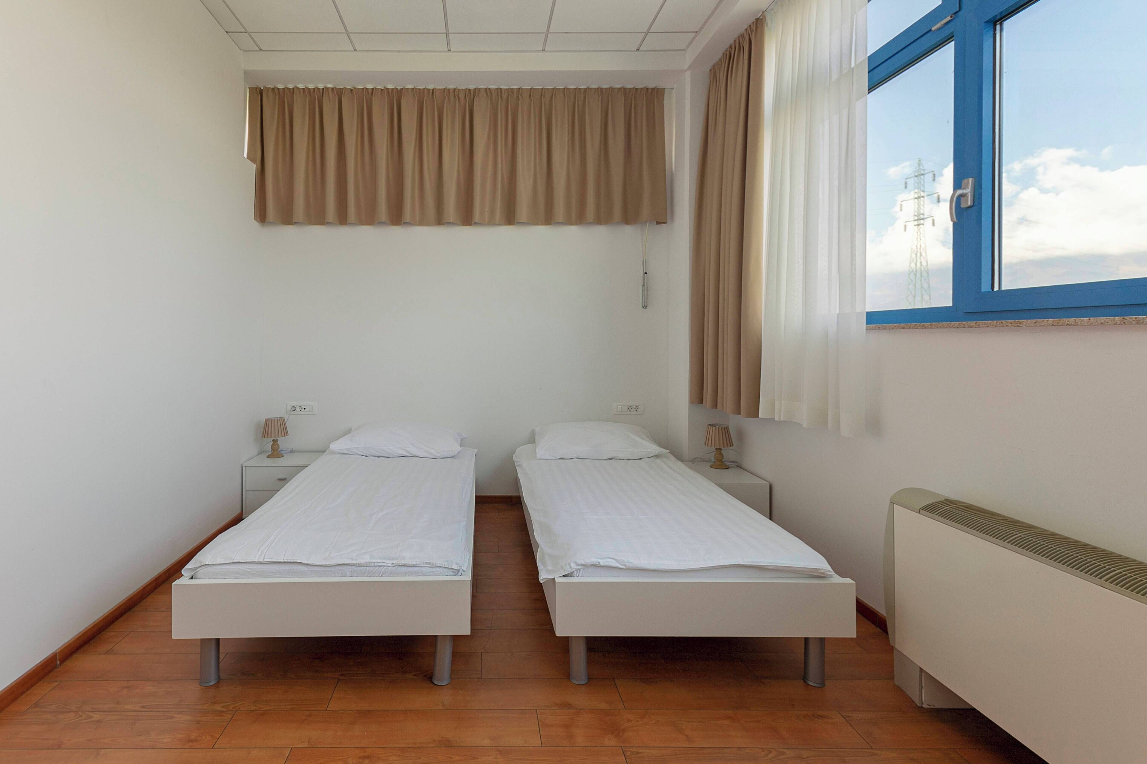 Une chambre avec deux lits simples, une fenêtre avec des rideaux, un radiateur et une prise électrique au mur.