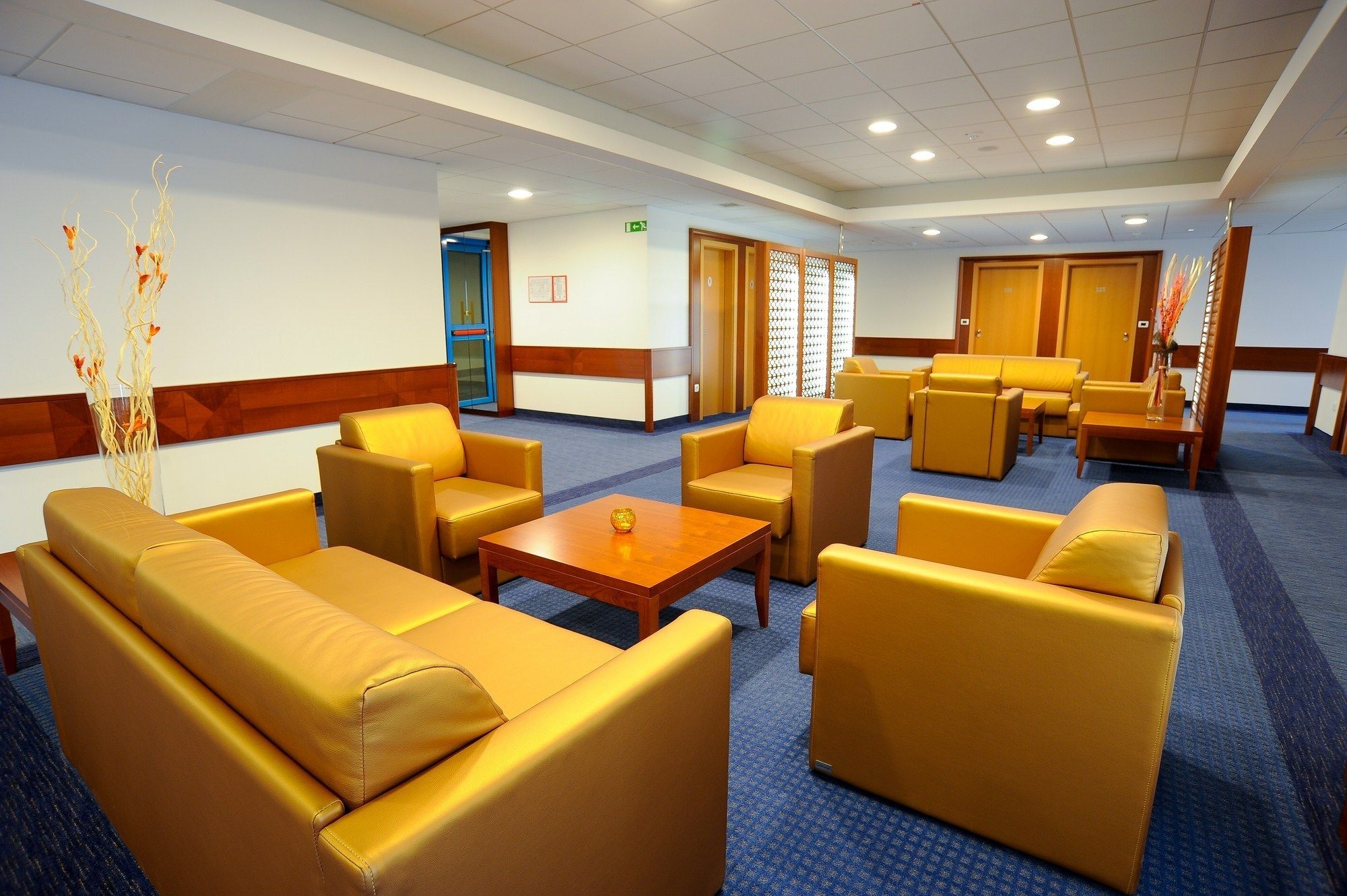Een hotellobby met gele banken en fauteuils, een houten salontafel en een blauw tapijt.
