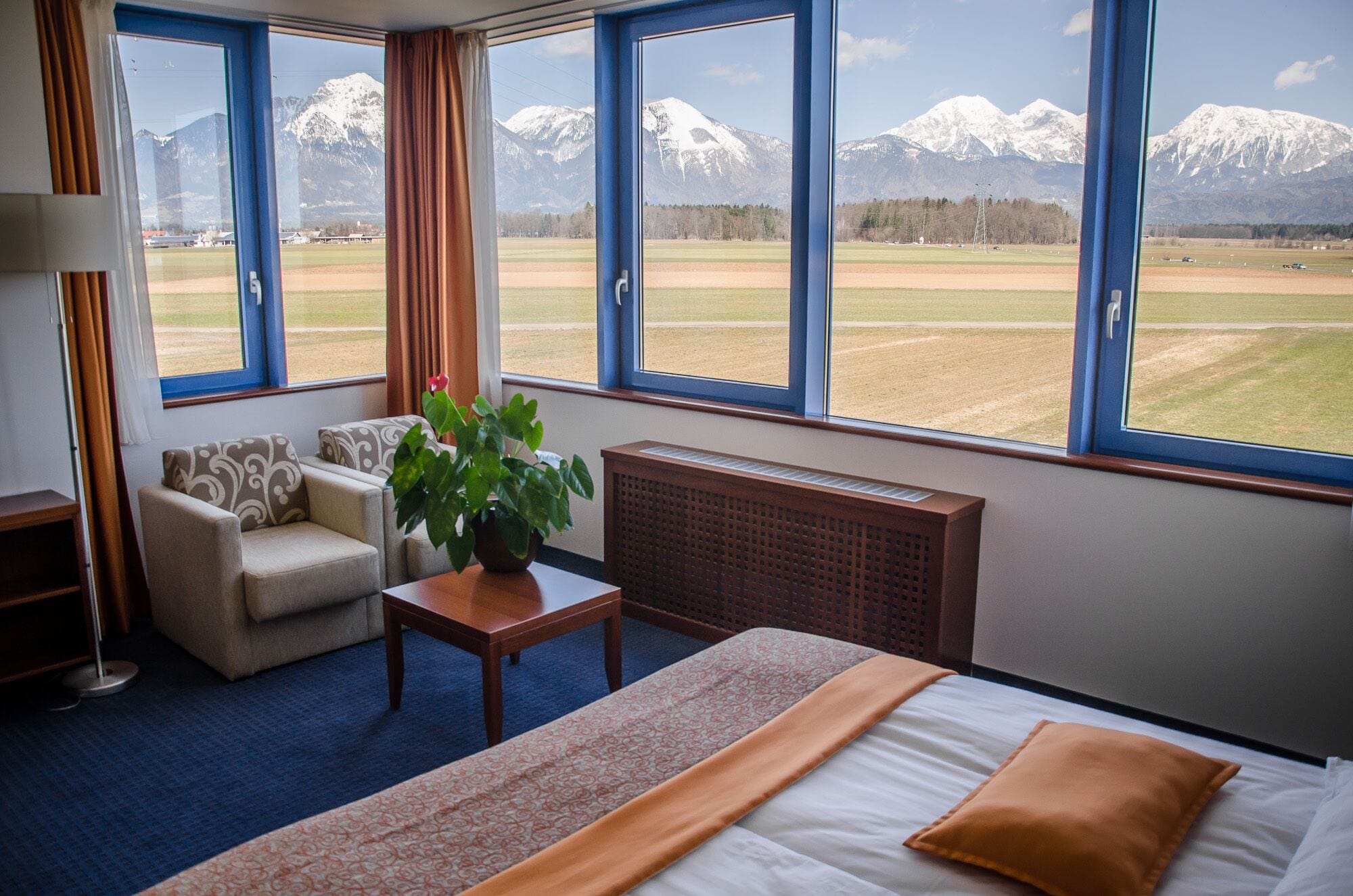 Een hotelkamer met een bed, een bank, een tafel met een plant en grote ramen met uitzicht op besneeuwde bergen.