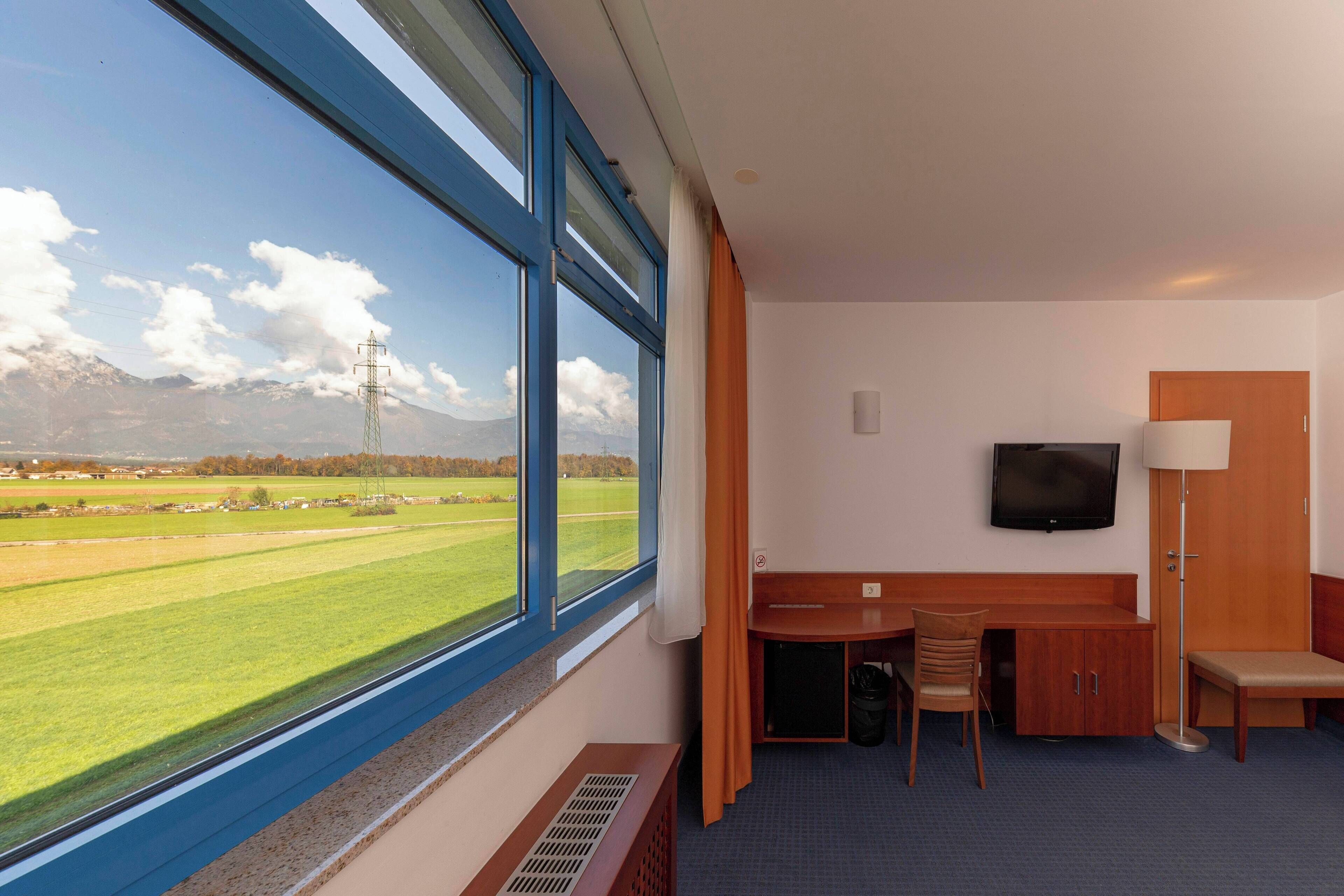 A hotel room with a large window, a desk, a chair, a TV, and a bench.