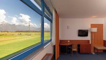 A hotel room with a large window, a desk, a chair, a TV, and a bench.