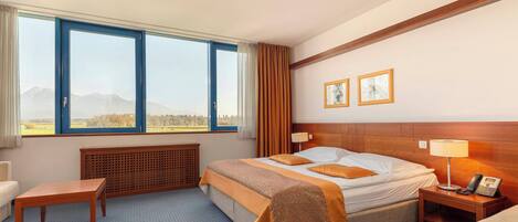 Habitación de hotel con una cama grande, una mesita de noche, una silla, un escritorio y vistas a la montaña desde la ventana.