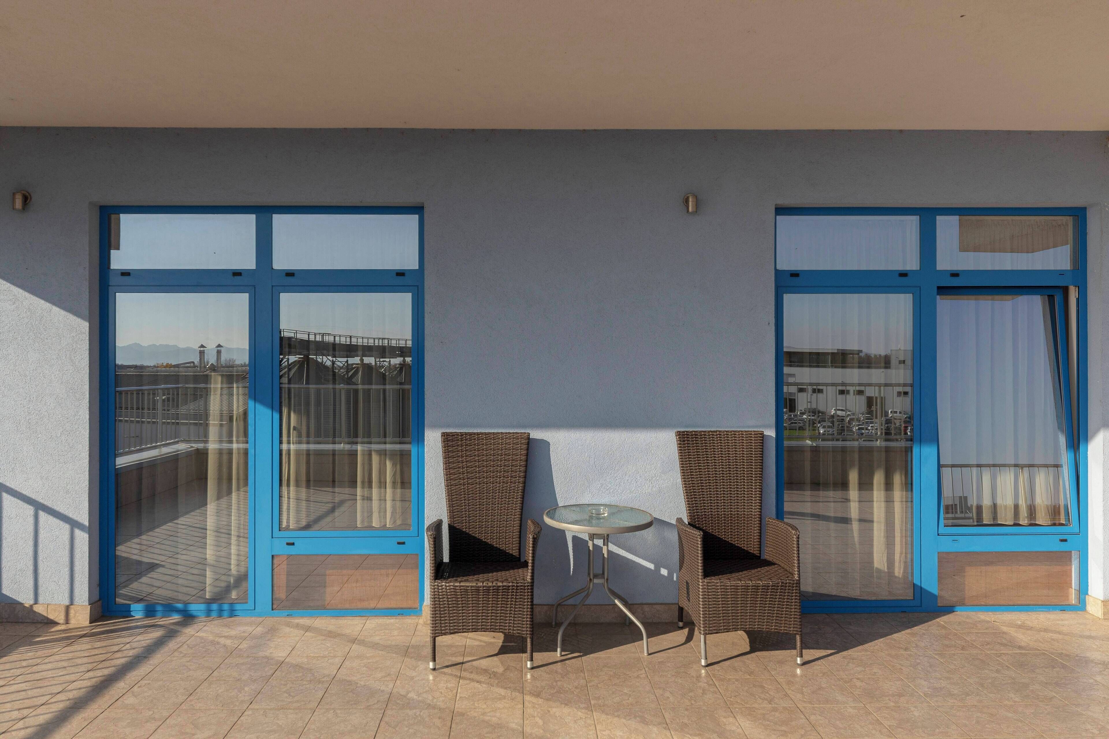 Un balcón con dos sillas de mimbre y una pequeña mesa de vidrio, ventanas grandes con marcos azules, y vistas a un puente y edificios.