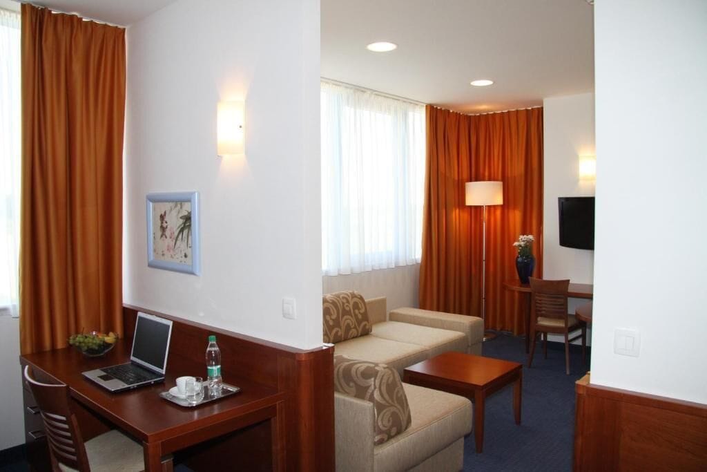A hotel room with a desk, laptop, and a sofa. There is a TV on the wall and a window with curtains.