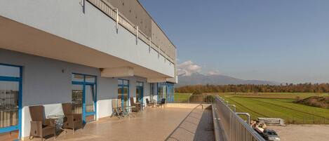 A building with a balcony, outdoor seating, and a view of a green field and mountains.