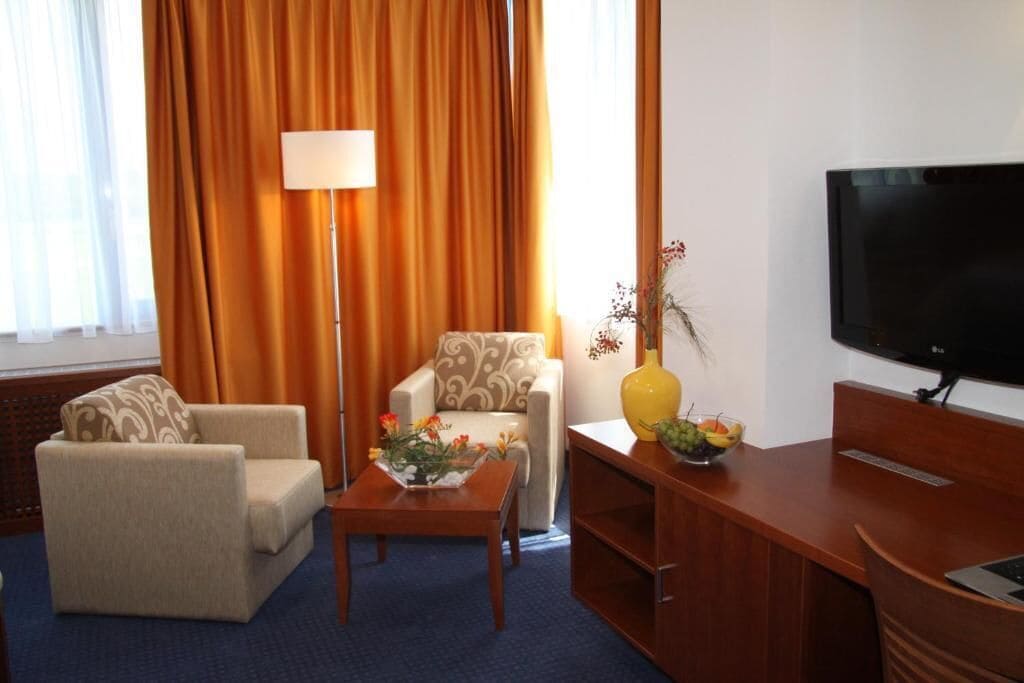 Habitación de hotel con sofá, sillón, mesa de centro, televisor y un florero con flores.