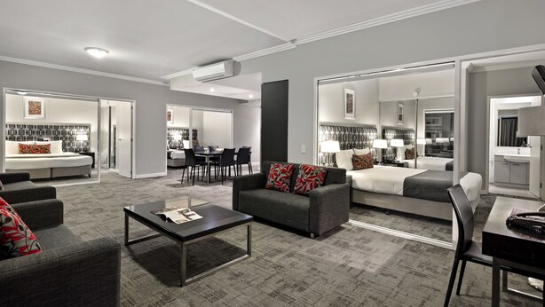 A modern hotel room with a grey sofa, a dining table, a separate bedroom with a bed, and a bathroom with a sink and mirror. - Quest Campbelltown (Campbelltown)
