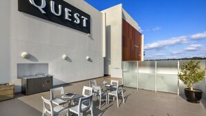 A modern outdoor patio area with a 'QUEST' sign, outdoor seating, a barbecue grill, and a potted plant. - Quest Campbelltown (Campbelltown)
