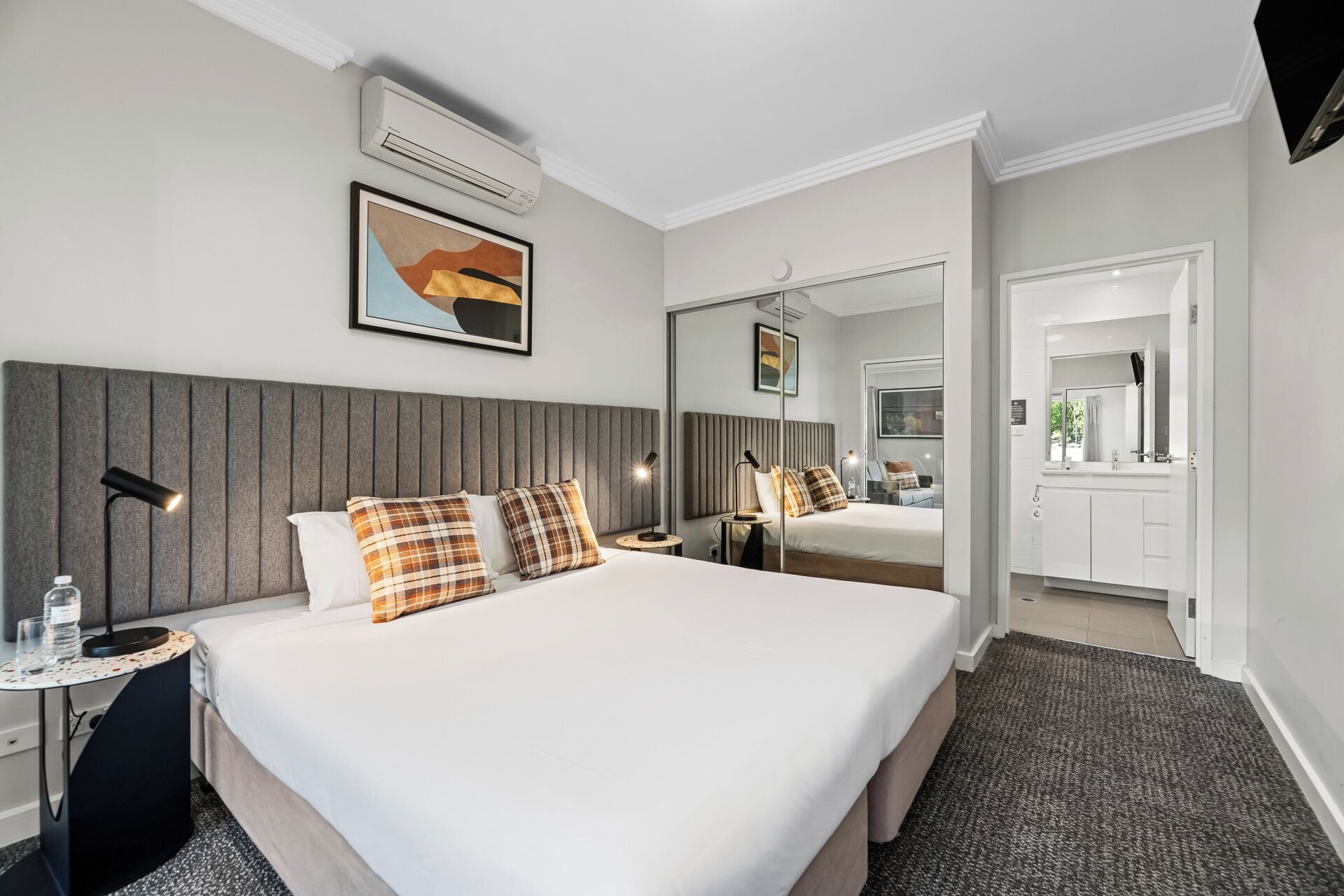 Una habitación de hotel moderna con una cama grande, una mesita de noche con lámpara, una botella de agua y un cuadro en la pared.