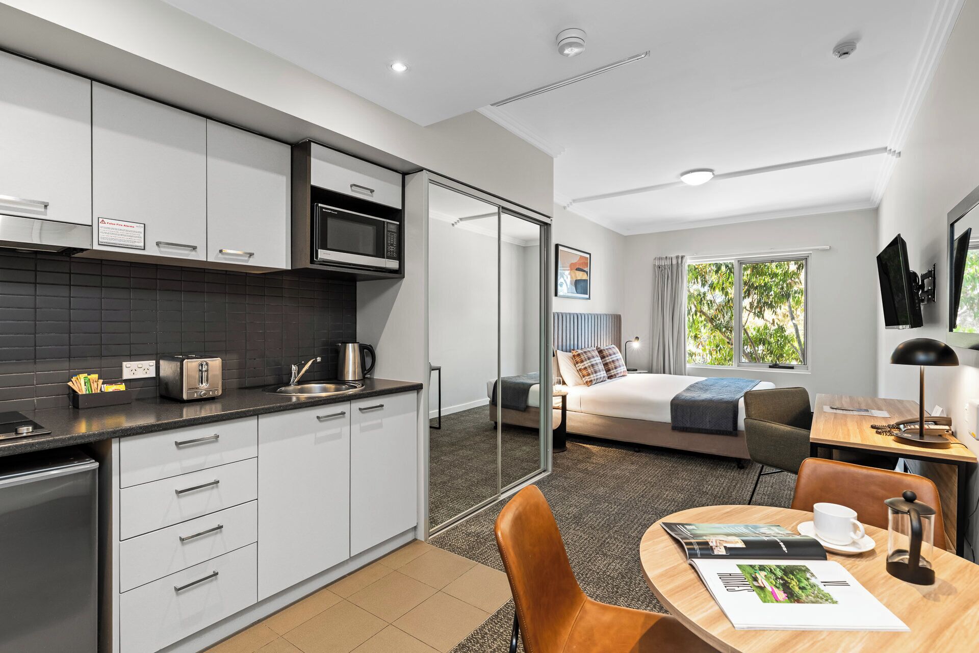 A modern hotel room with a kitchenette, a bed with a view, a dining area, and a desk.