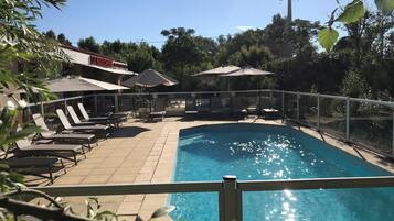 Une piscine entourée d’une clôture et d’arbres, avec des chaises longues et des parasols à proximité.