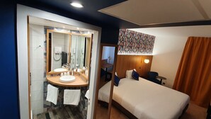 A hotel room with a double bed, a large mirror, two sinks, a bedside table, and a small table with a chair.