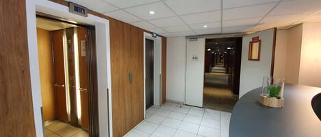 Un hall d’hôtel avec un ascenseur, un comptoir d’accueil et un couloir menant aux autres chambres.