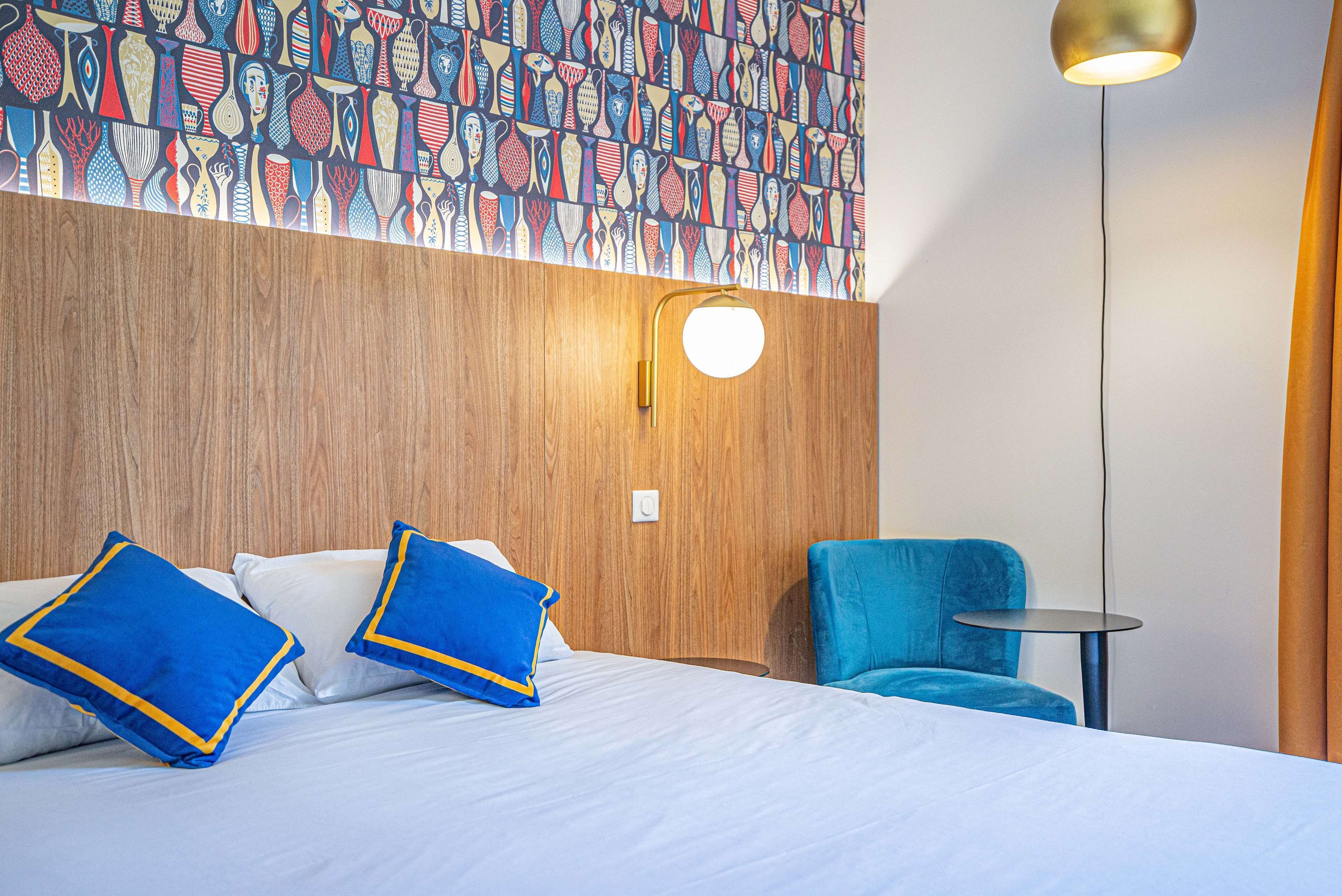 A hotel room with a wooden headboard, a bed with white linens and blue pillows, a blue armchair, a small round table, and a wall with a colorful abstract pattern.