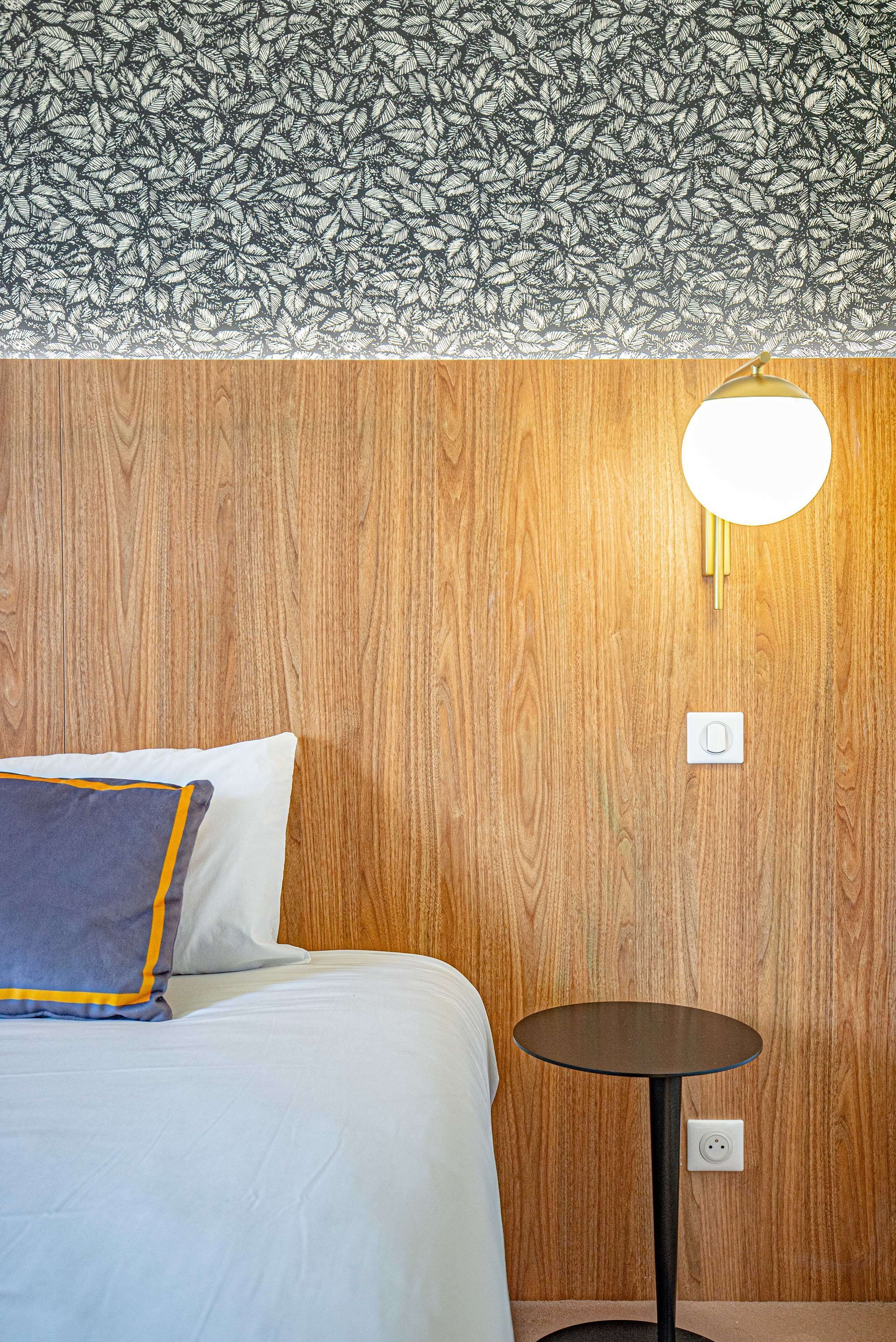 A hotel room with a wooden wall, a round table with a light, and a bed with a pillow.