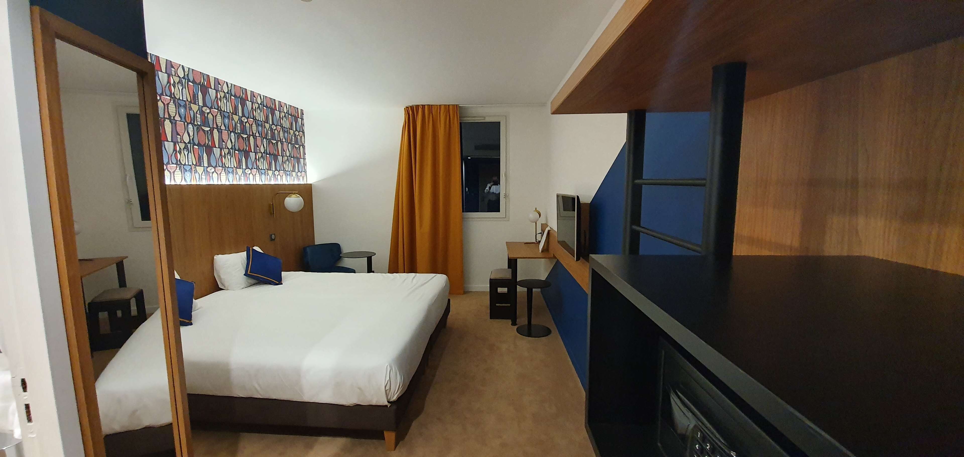 A hotel room with a bed, a desk, a chair, a small table, a TV, and a mirror.