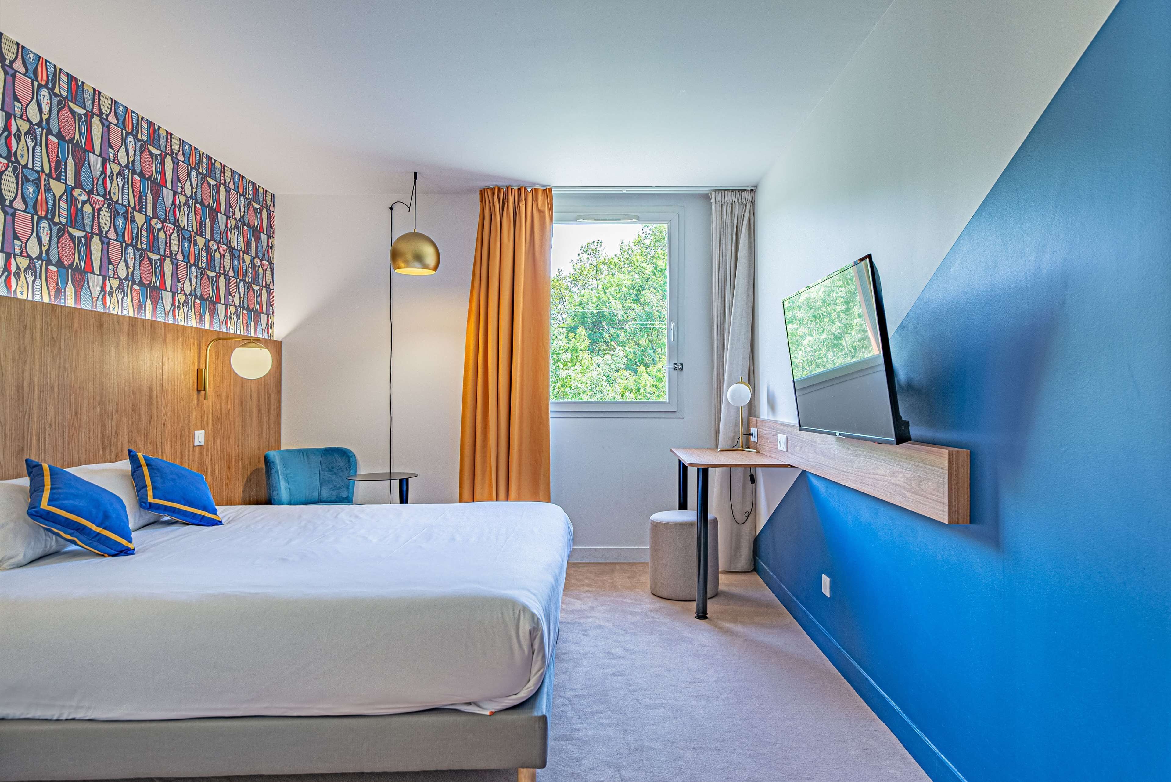 A modern hotel room with a large bed, a desk, a television, and a view of greenery outside.