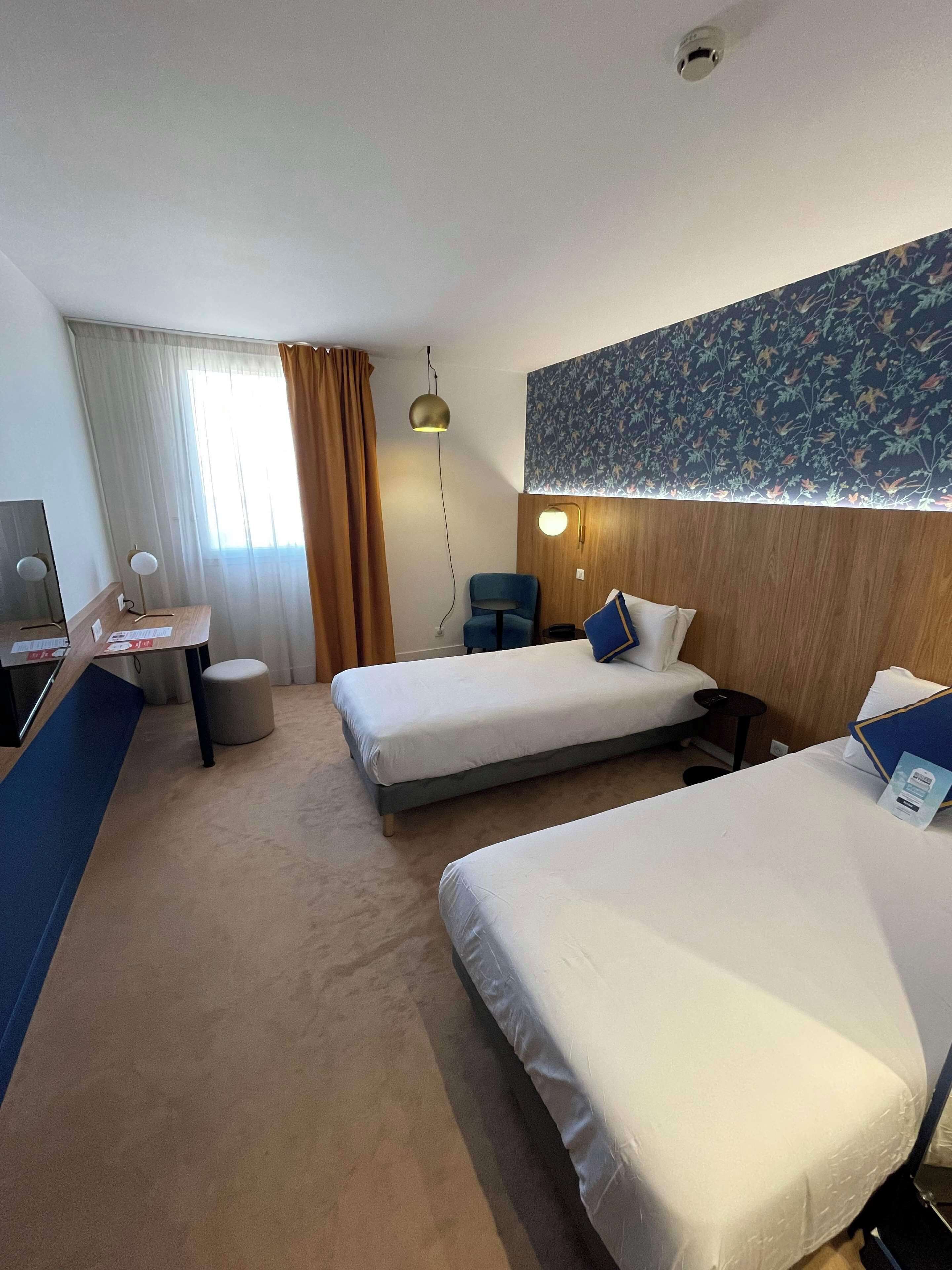 A hotel room with two beds, a desk, a chair, a small round ottoman, and a wall-mounted lamp.