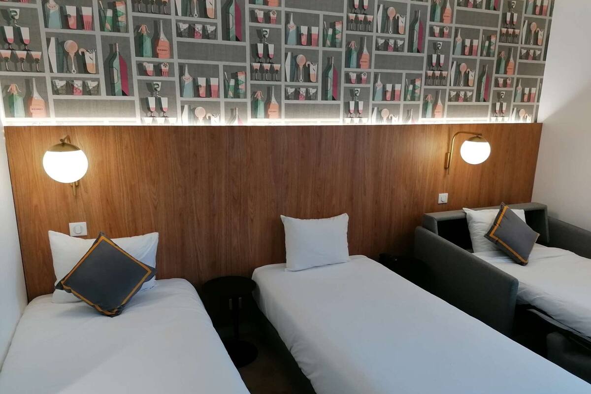 A hotel room with three single beds, each with a pillow and a small bedside table. The room has wooden paneling on the walls and a wall-mounted light fixture above the beds.