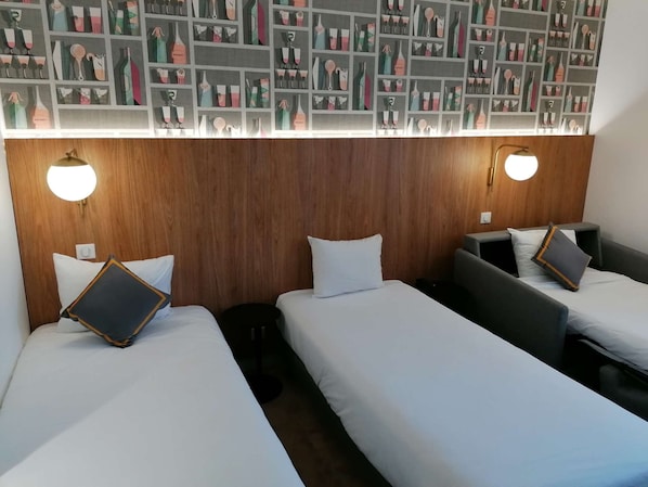 Une chambre d’hôtel avec trois lits simples, chacun équipé d’un oreiller et d’une petite table de chevet. Les murs de la chambre sont recouverts de lambris en bois, et un luminaire est fixé au mur au-dessus des lits.